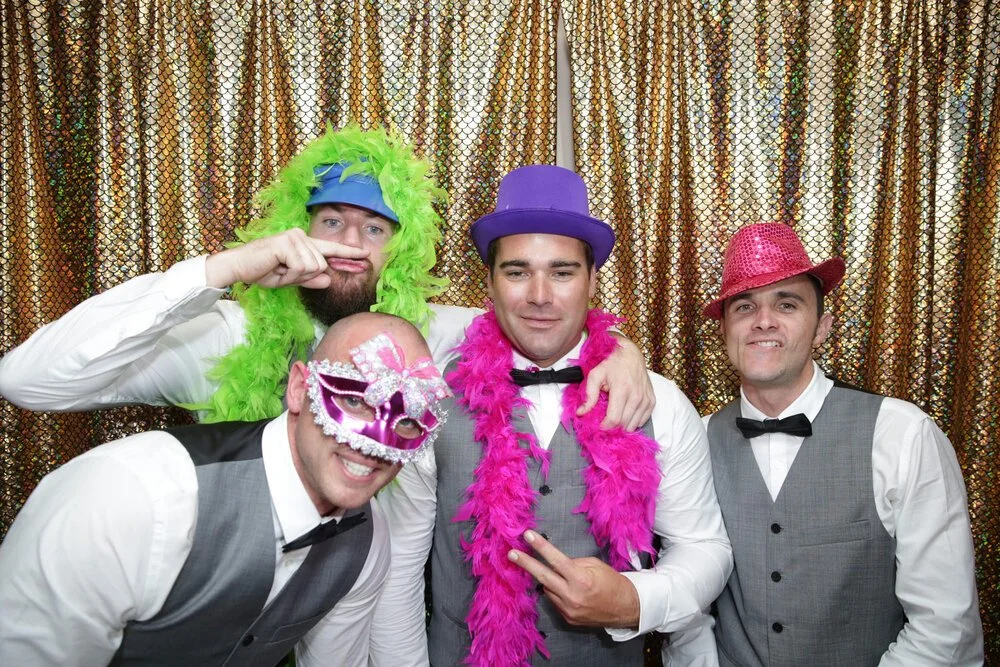 Four men in formal wear with colorful hats and feather boas posing in front of a shiny backdrop.