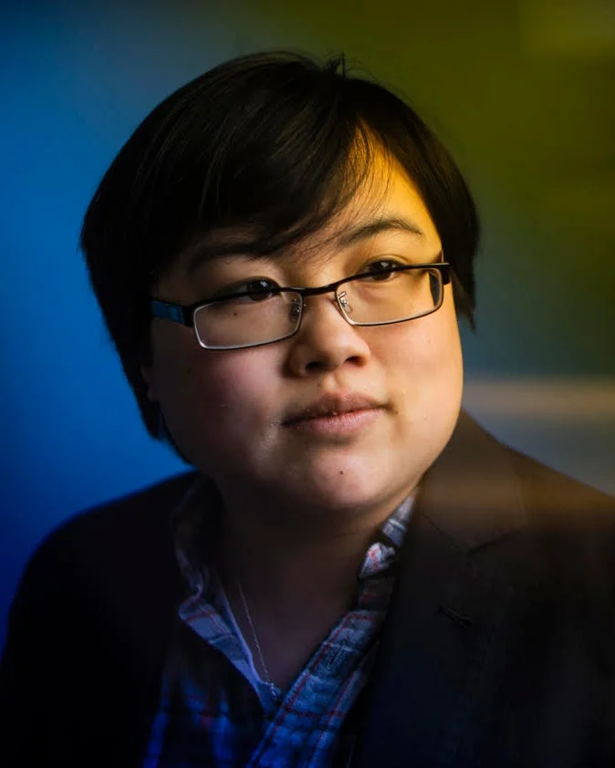 Photo: Headshot of Lydia Brown, young East Asian person, with stylized blue and yellow dramatic background. They are looking in the distance and wearing a plaid shirt and black jacket. Photo by Adam Glanzman.