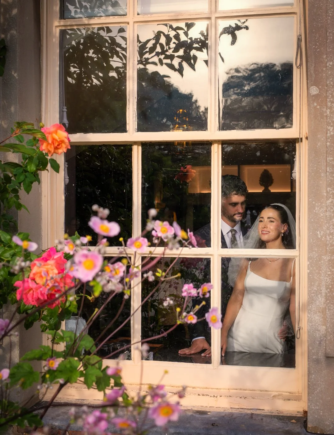 A bride and groom inside a house, seen through a window with multiple panes, with pink and orange flowers outside.