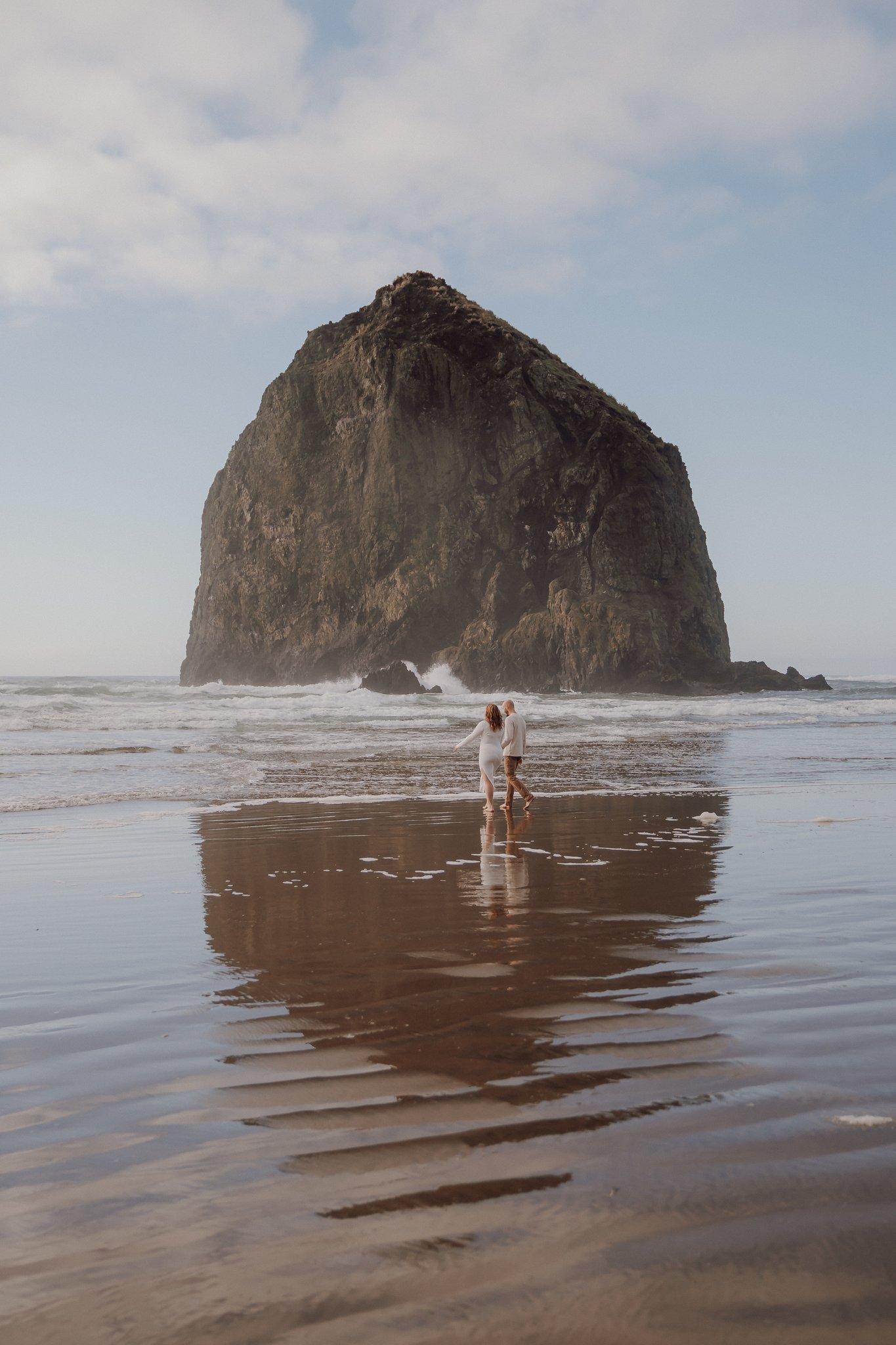 A couple walking on a sandy beach towards a large rock formation with waves in the background, under a partly cloudy sky.