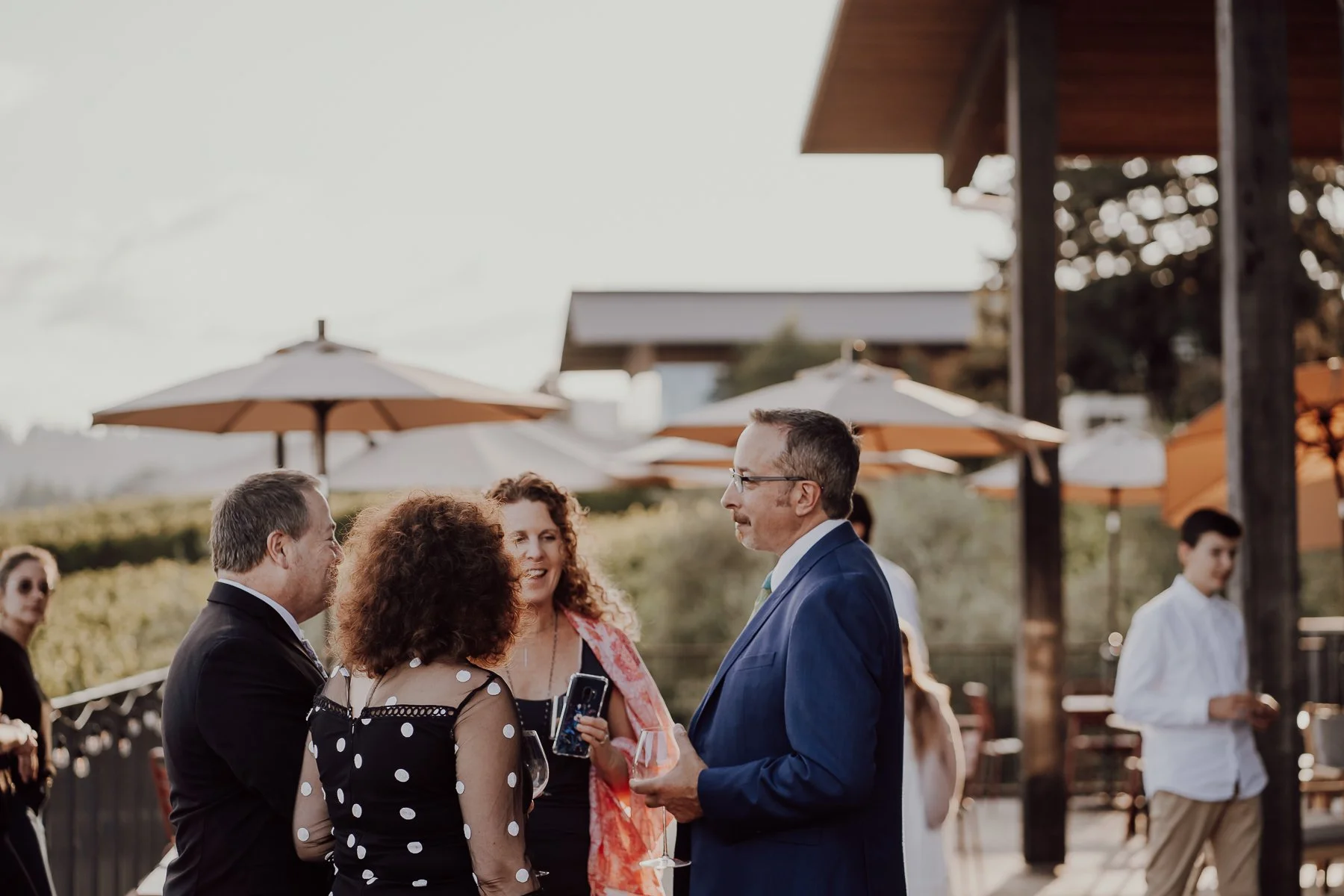 Group of people socializing outdoors under patio umbrellas, dressed in formal attire, with one person wearing a polka dot dress, another in a blue suit, and holding drinks.