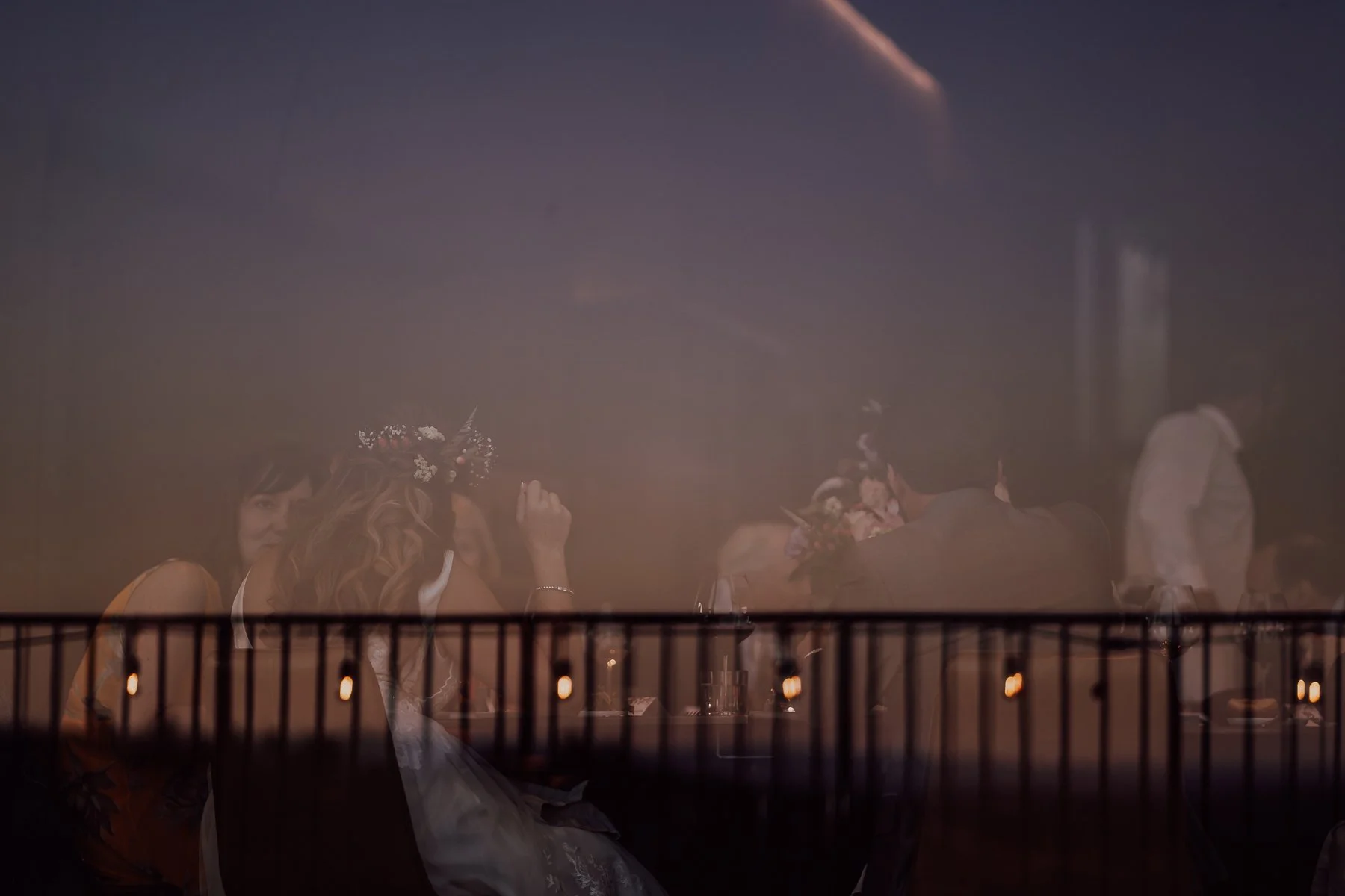 Reflection showing people dining indoors with soft lighting, blurred view of a railing and small lights outside.