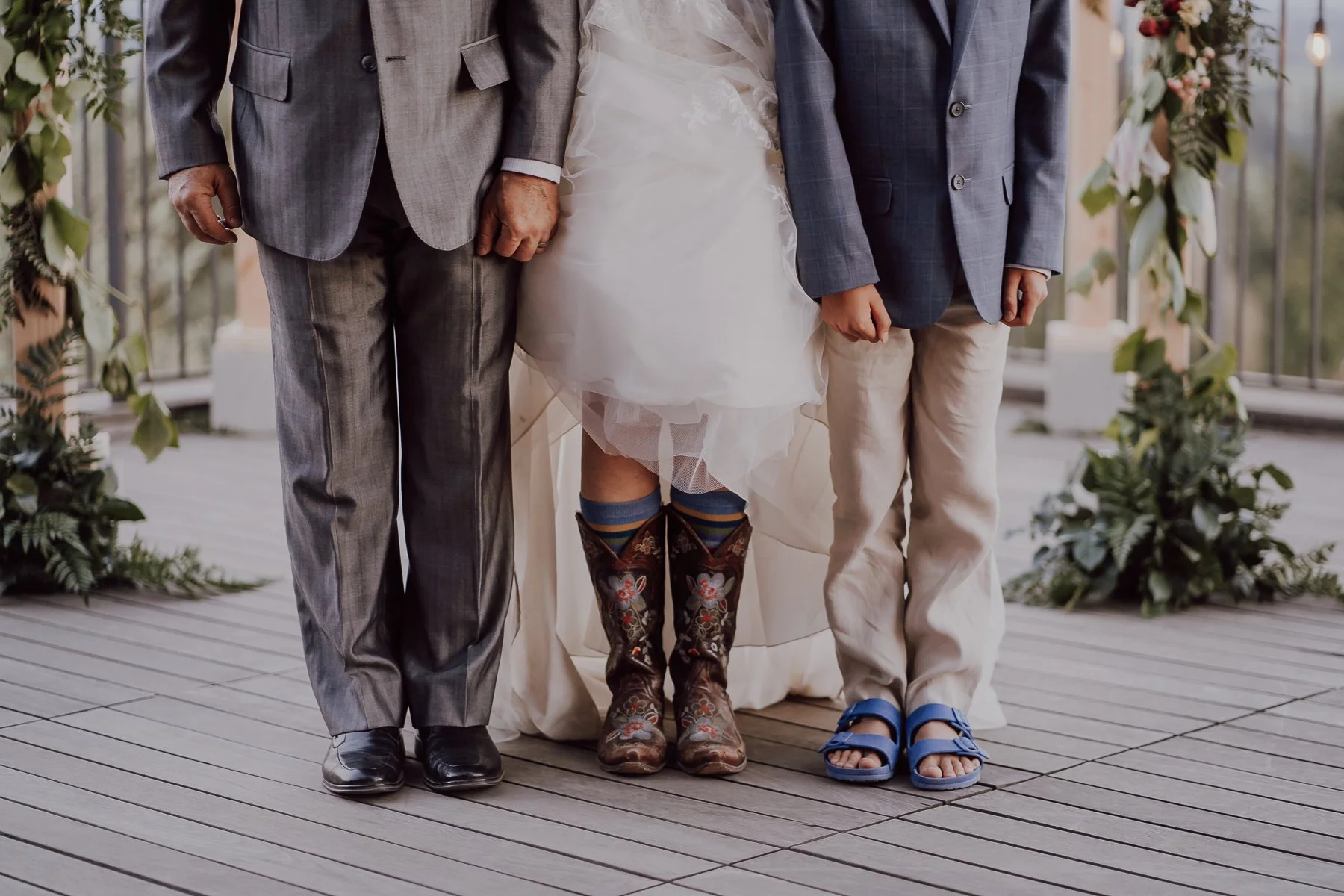 Three people standing; an adult in a gray suit and black shoes, a person in a wedding dress with embroidered cowboy boots, and a child in a blazer and beige pants with blue sandals, on a wooden deck with greenery.