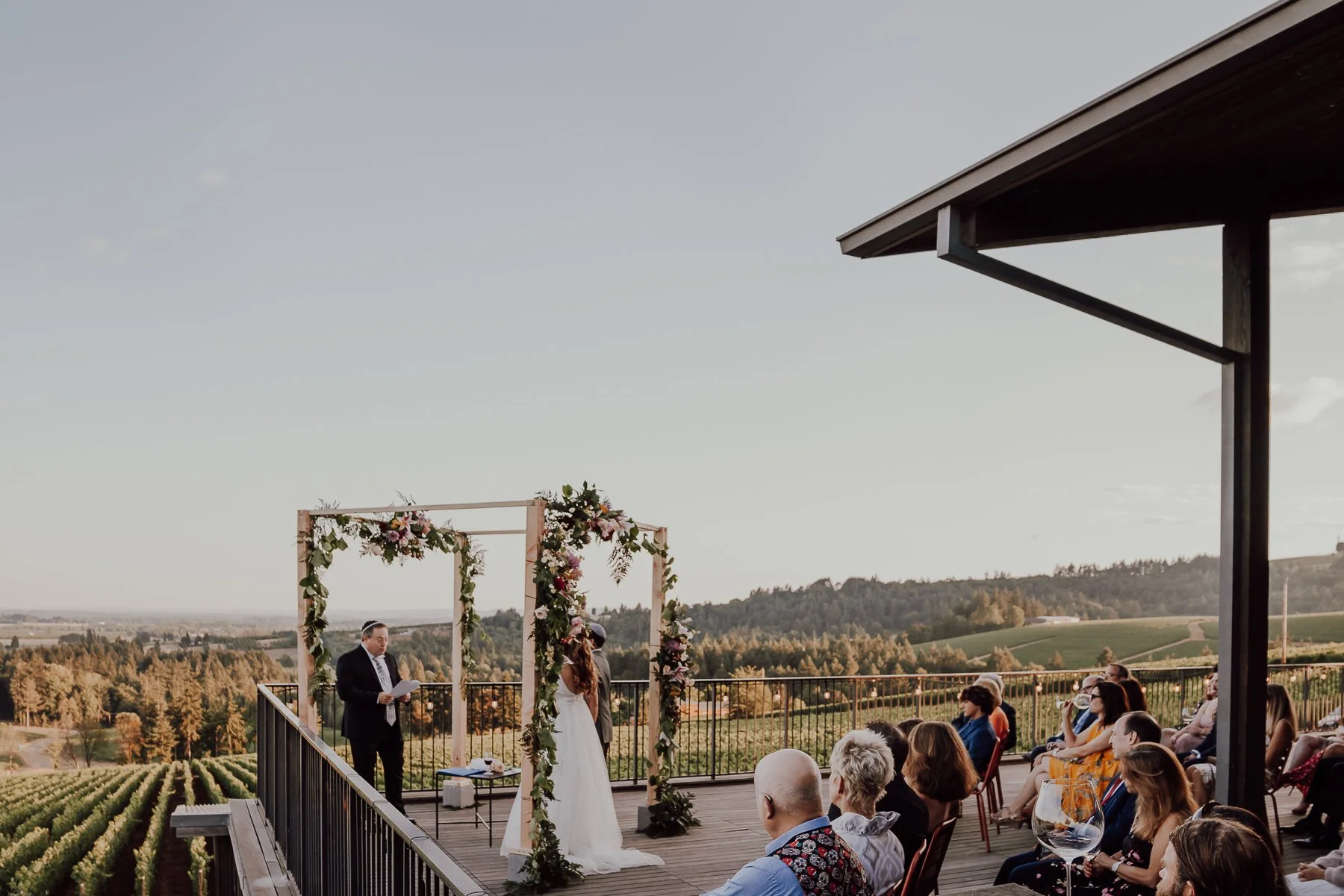 Outdoor wedding ceremony on a deck overlooking a vineyard, featuring a floral arch and seated guests.