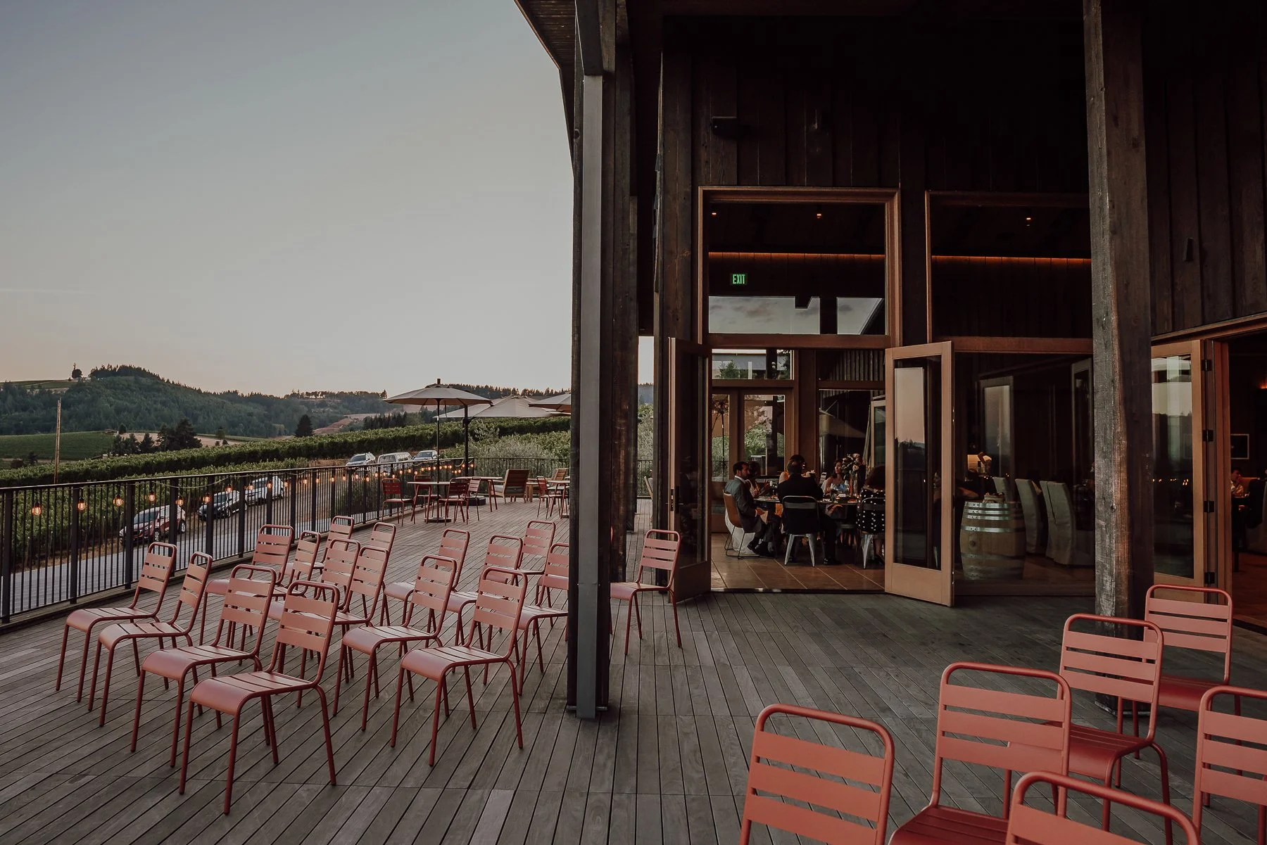 Outdoor patio with red chairs, overlooking vineyard, dining area indoors, evening setting, umbrellas, and scenic landscape.