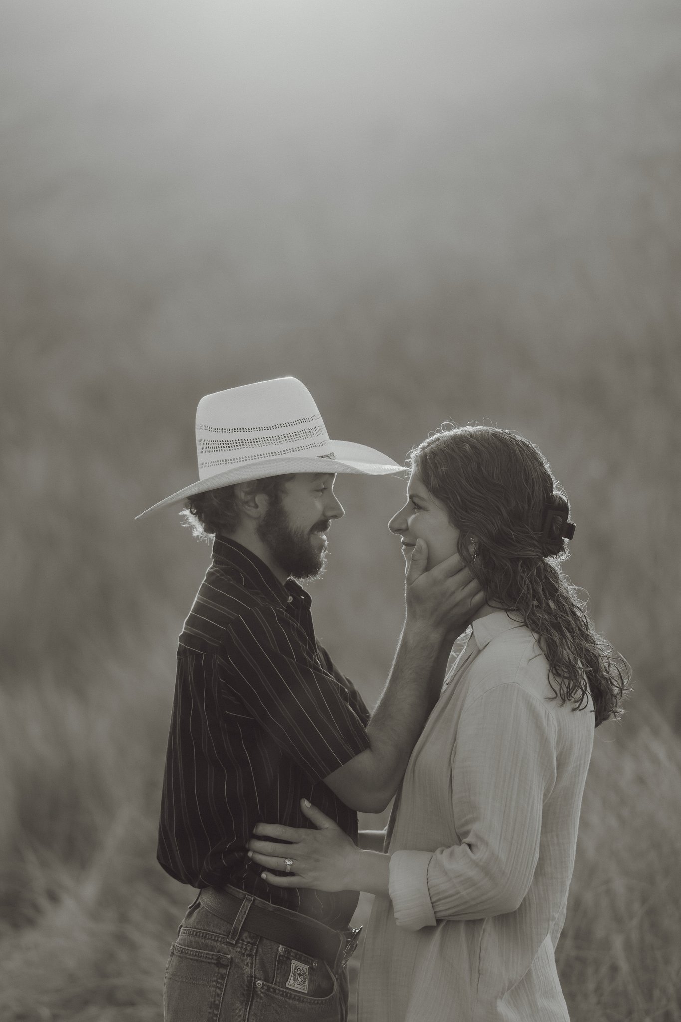 A monochrome image of a couple in a field. The man is wearing a cowboy hat and a striped shirt, gently touching the woman's face. She is wearing a light-colored shirt and has her hair tied back, with her hand resting on his chest.