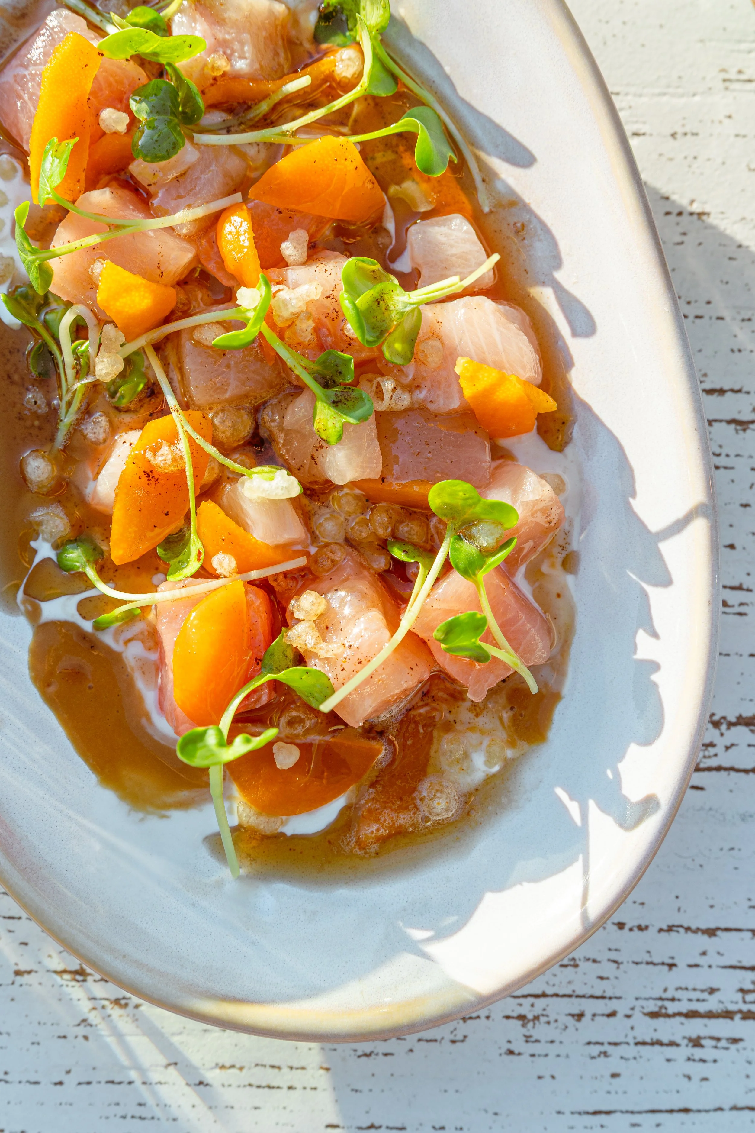 A dish of salmon tartare topped with microgreens, diced yellow and orange vegetables, and a sauce, served on a white plate on a rustic wooden surface.
