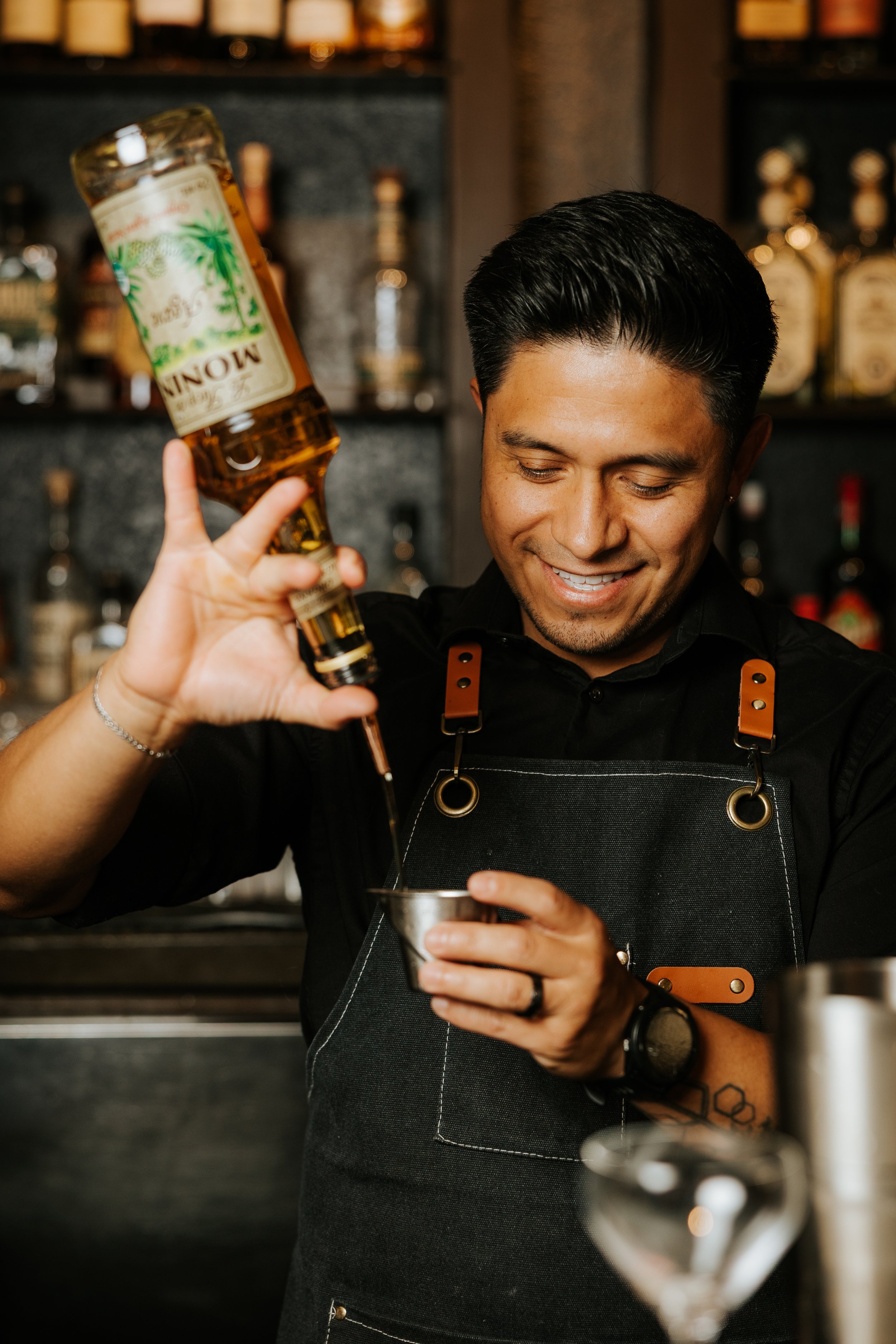 A bartender with dark hair, wearing a black shirt and apron, pouring a drink into a cocktail shaker in a bar with liquor bottles in the background.