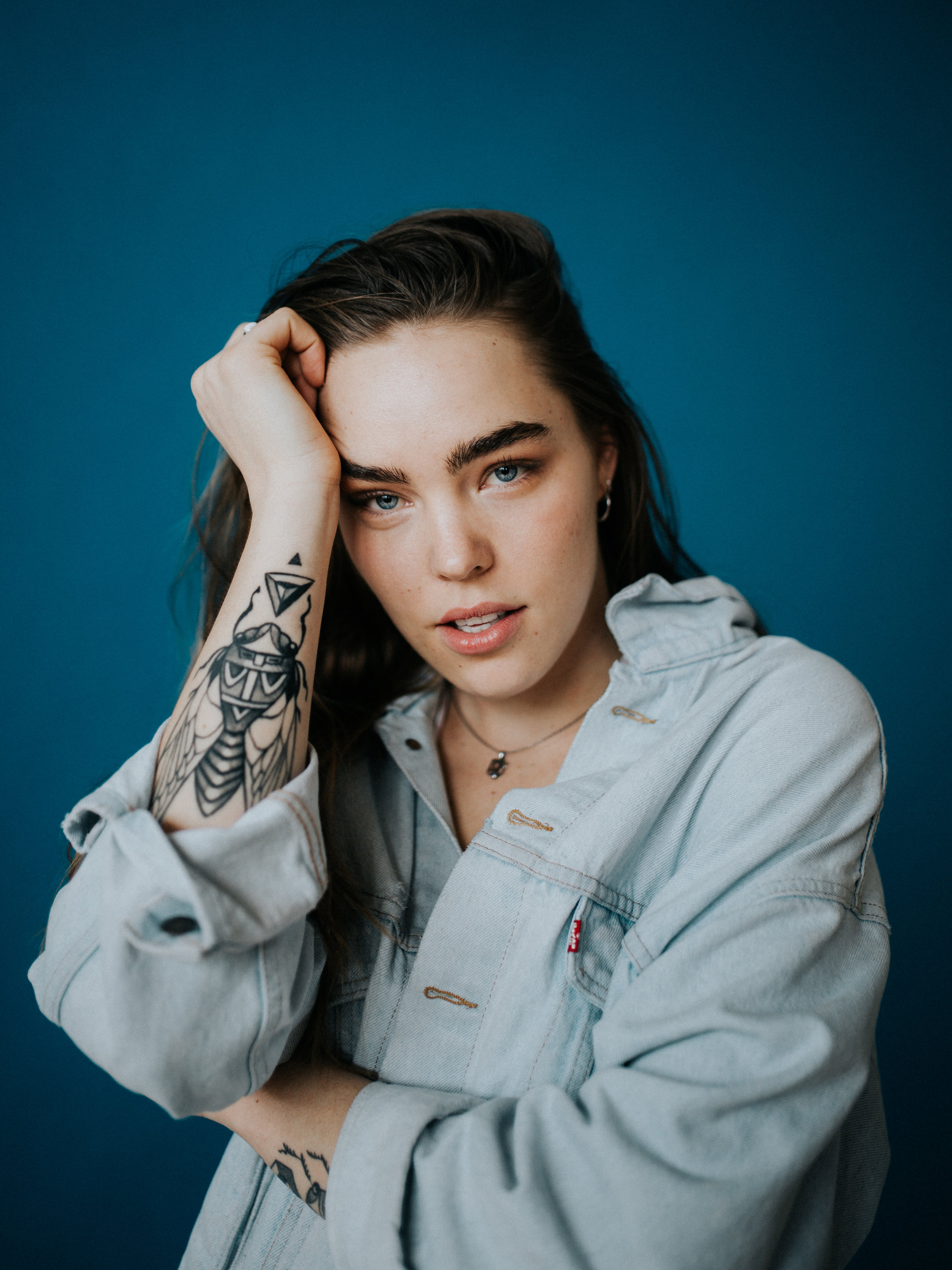 A woman with dark hair and blue eyes posing against a solid blue background. She is wearing a light-colored denim jacket and has tattoos on her left arm.