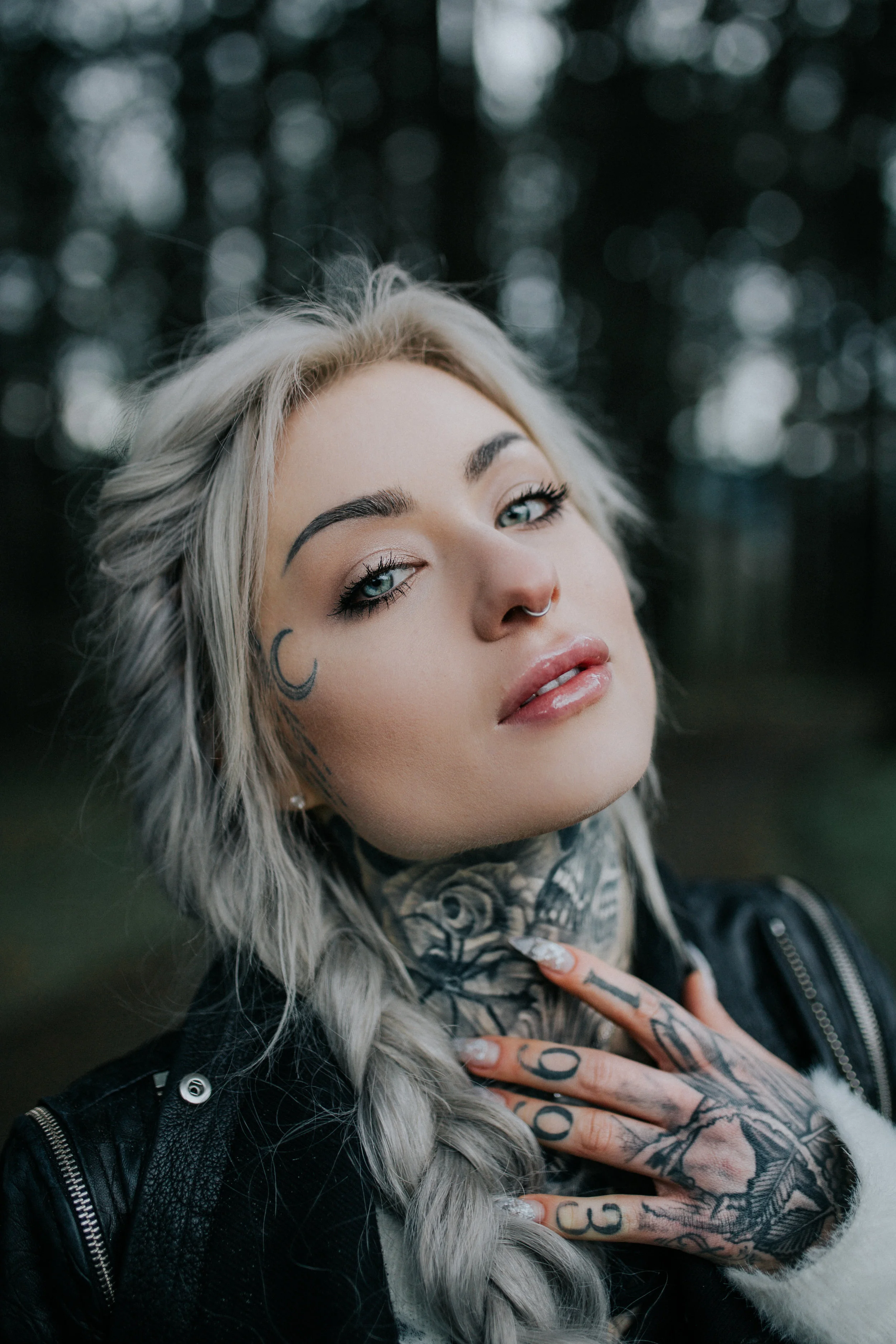 A young woman with platinum blonde hair, tattoos on her face and hands, wearing a septum piercing, makeup, and a black leather jacket.