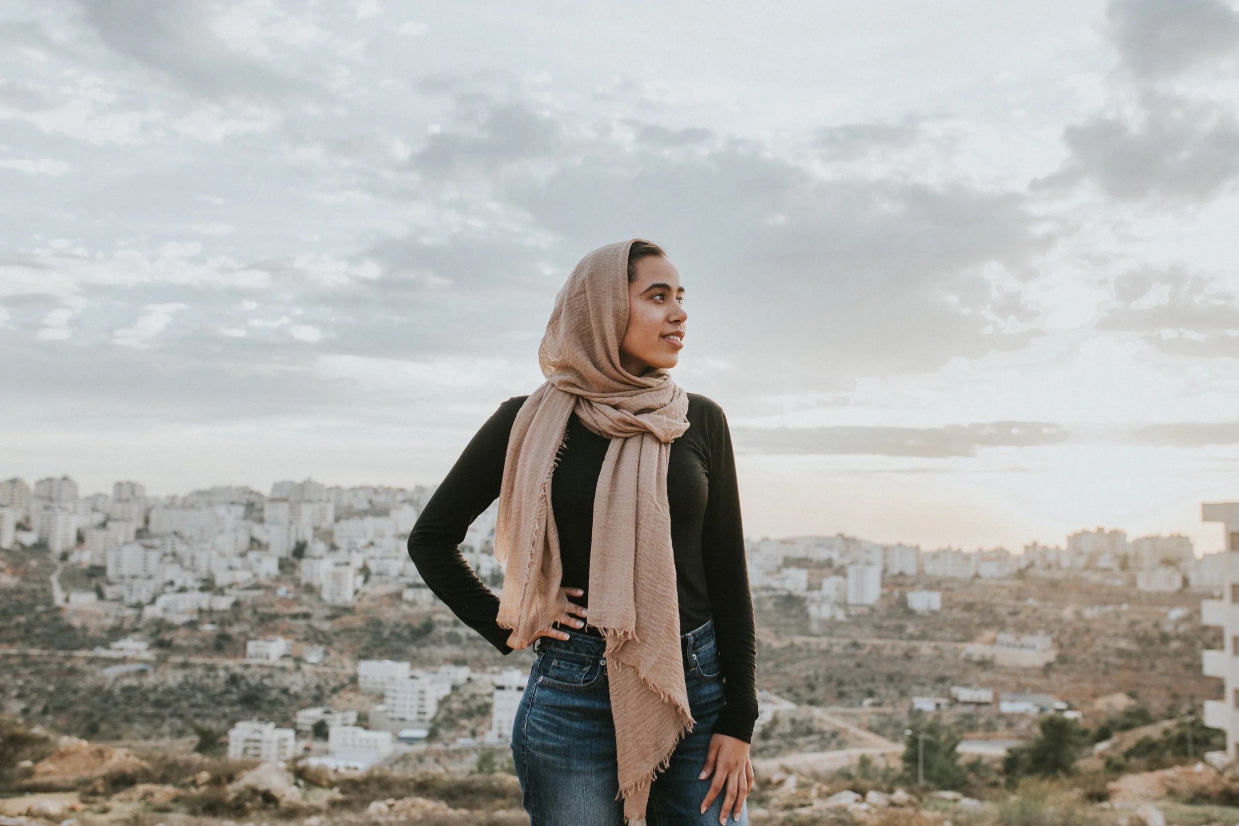 A woman wearing a tan hijab and black shirt stands outdoors overlooking a Ramallah, Palestine in the West, during cloudy weather.