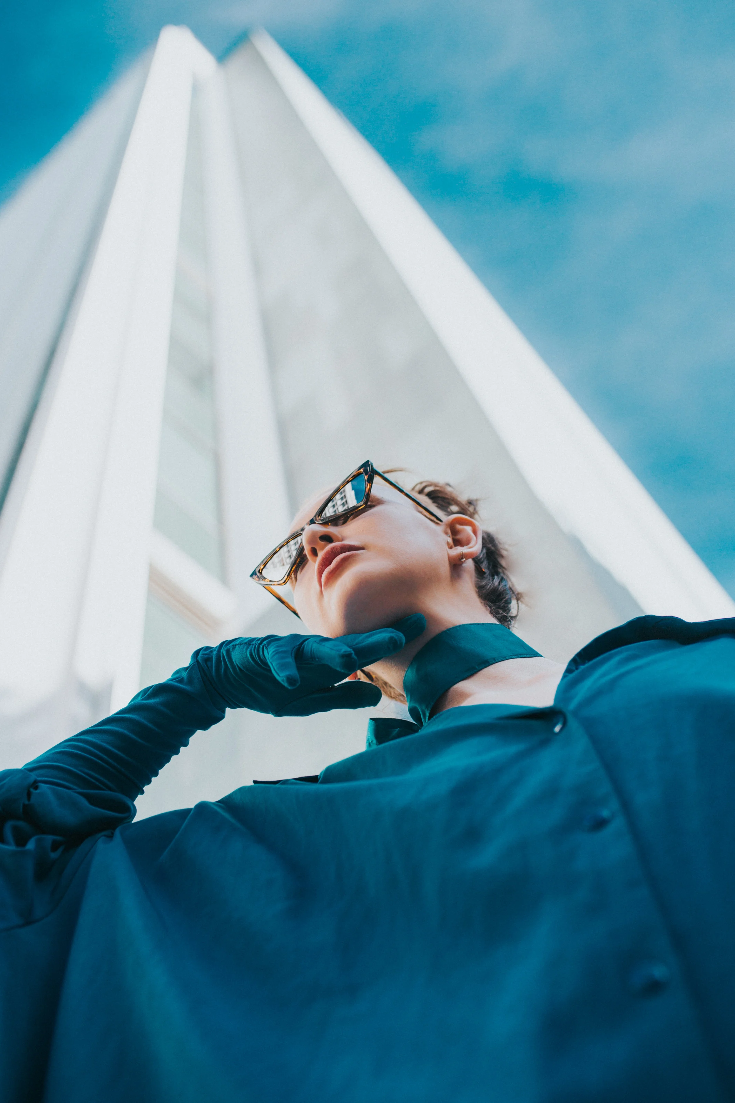 A woman in a dark blue jacket and black gloves is adjusting her glasses while standing outdoors under a blue sky and modern white building.