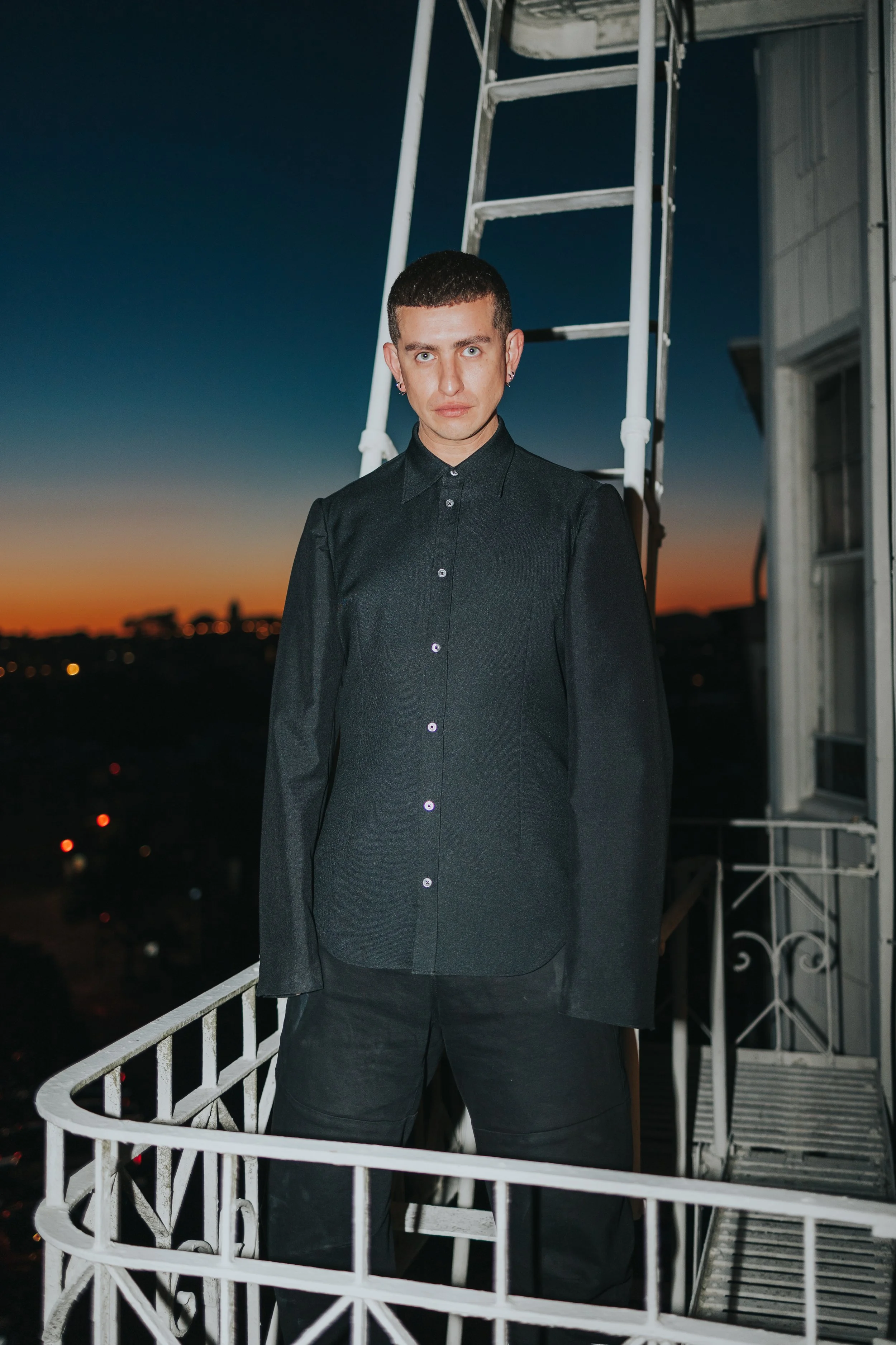 A person with short dark hair, wearing earrings and a black button-up shirt, standing on a balcony at sunset with buildings in the distance. There is a white metal railing and a ladder in the background.