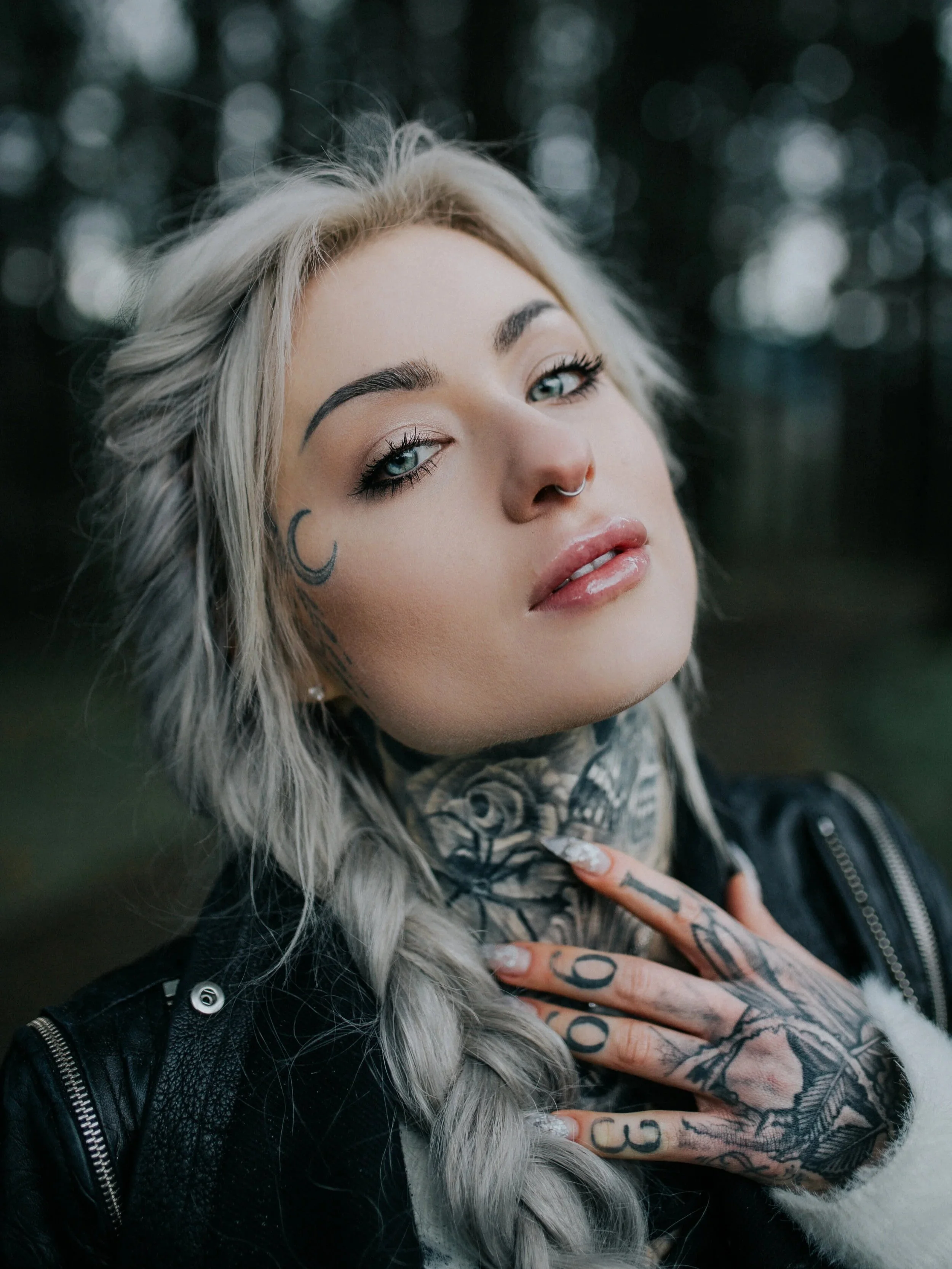 A young woman with platinum blonde hair in a braid, tattoos on her neck and hands, and piercings, posing outdoors with a blurred forest background.