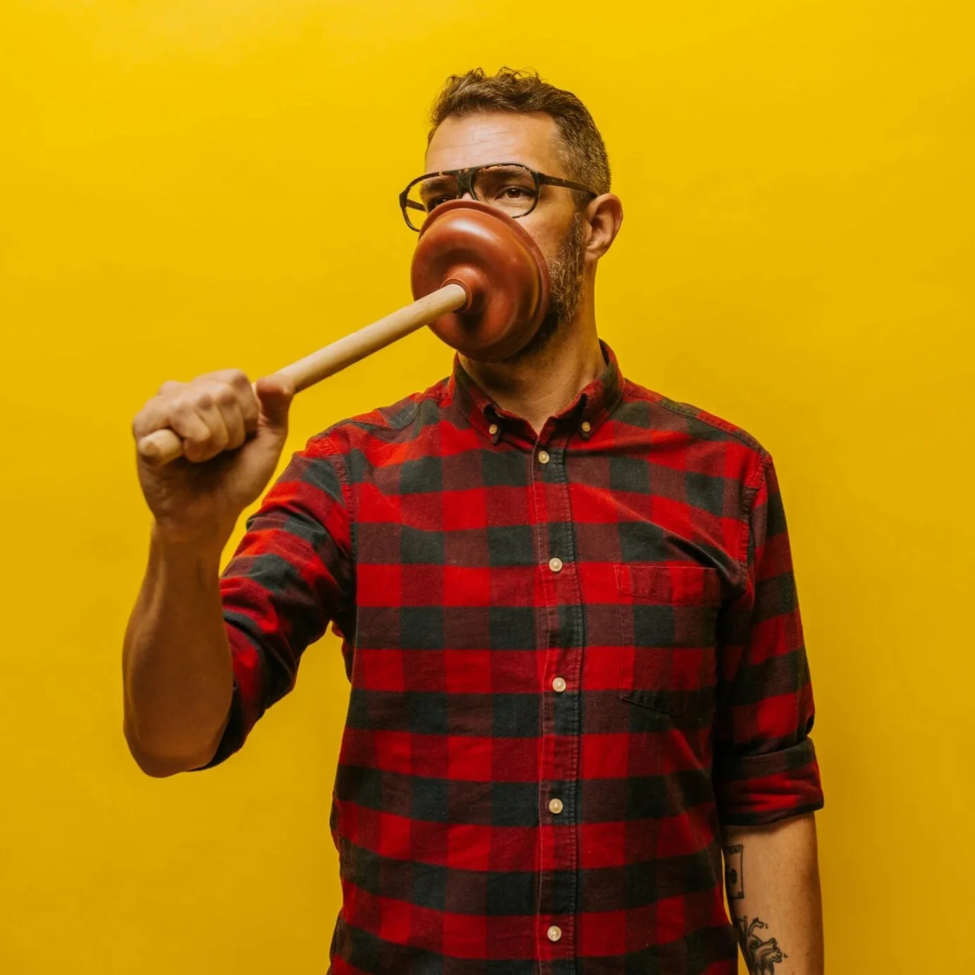 A man in glasses and a red plaid shirt holding a plunger against his face, standing against a yellow background.