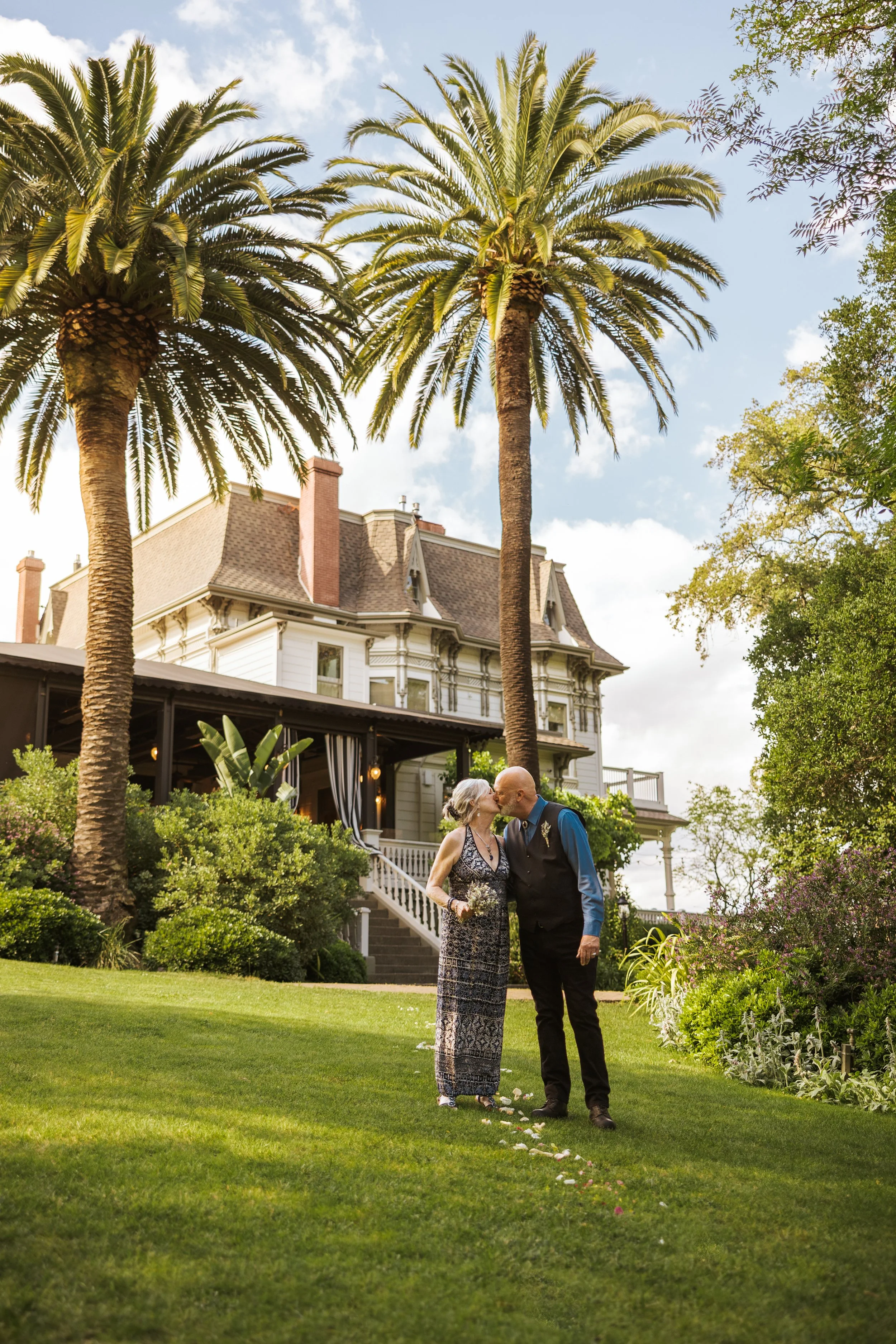 An elderly couple sharing a kiss in a lush garden in front of a Victorian-style house with palm trees.