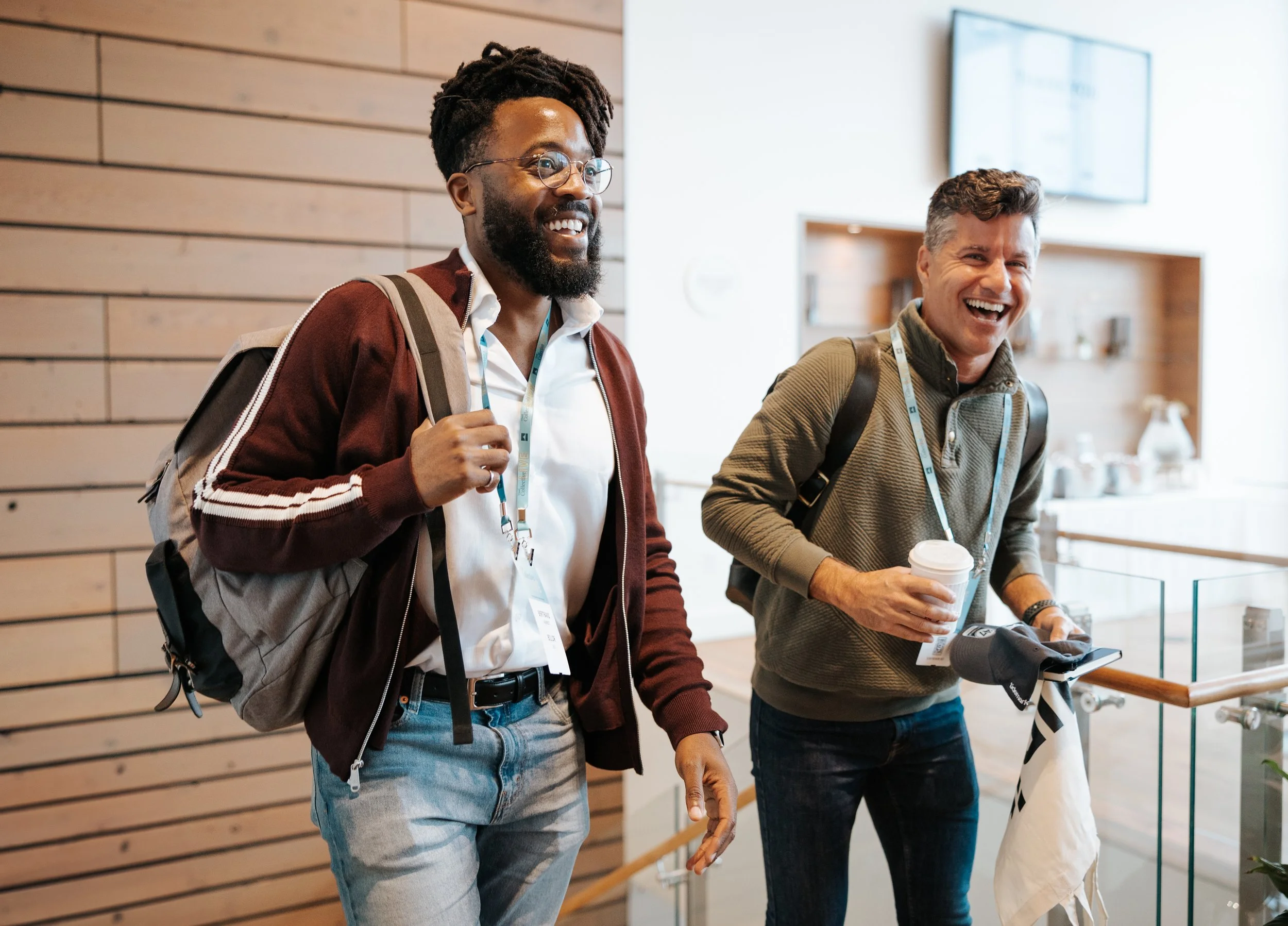 Two men smiling and walking with backpacks in a modern indoor setting, one holding a coffee cup.