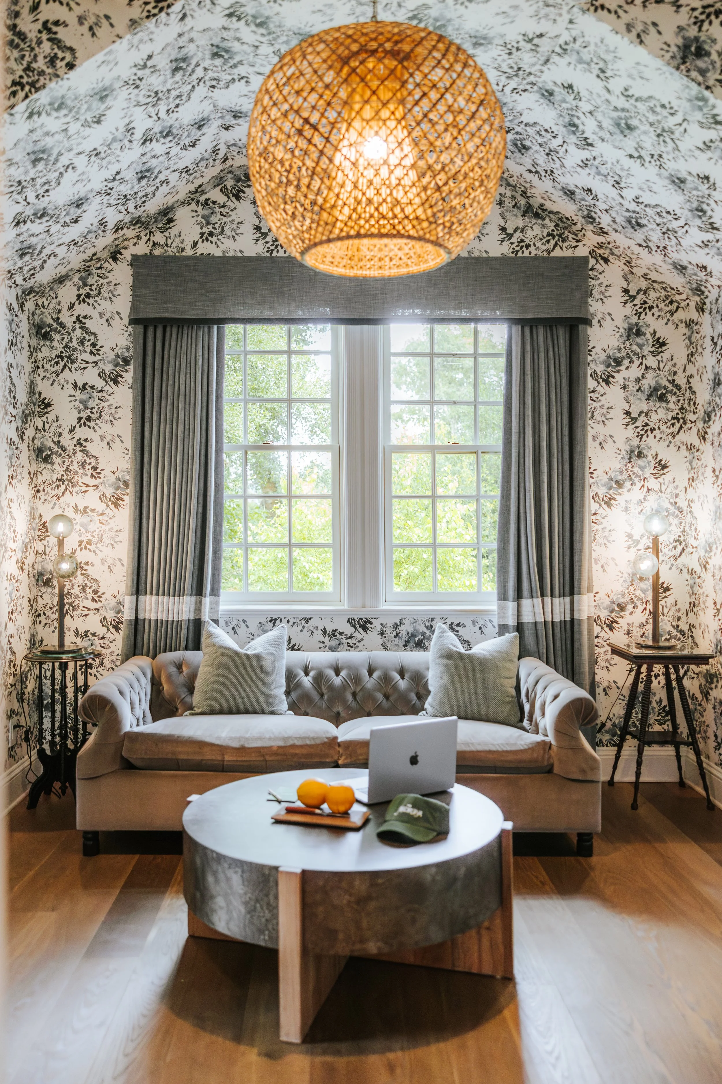 Living room with a tufted beige sofa, white pillows, a coffee table with oranges and a laptop, large window with curtains, floral wallpaper ceiling, wicker pendant light, and side lamps.