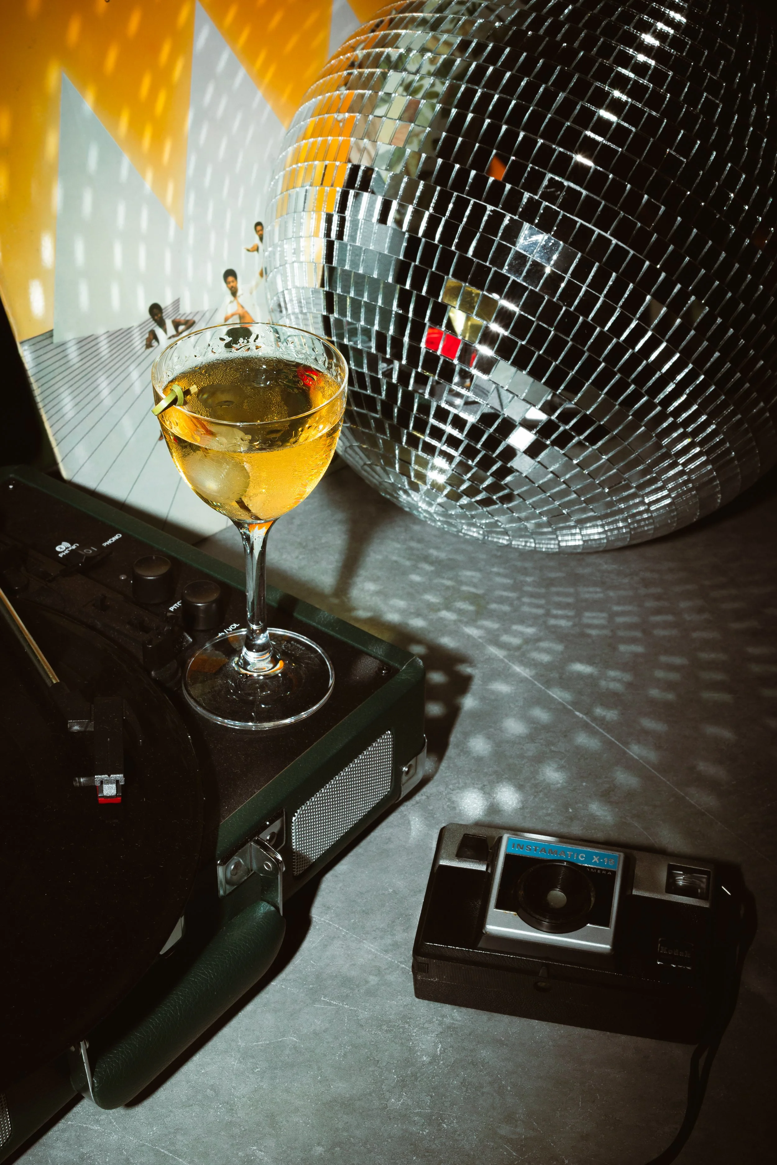 A disco ball with mirrored tiles reflecting light, a glass of mixed drink with fruit garnish on a DJ turntable, a vintage camera on a dark surface, and a magazine showing images of dancers.
