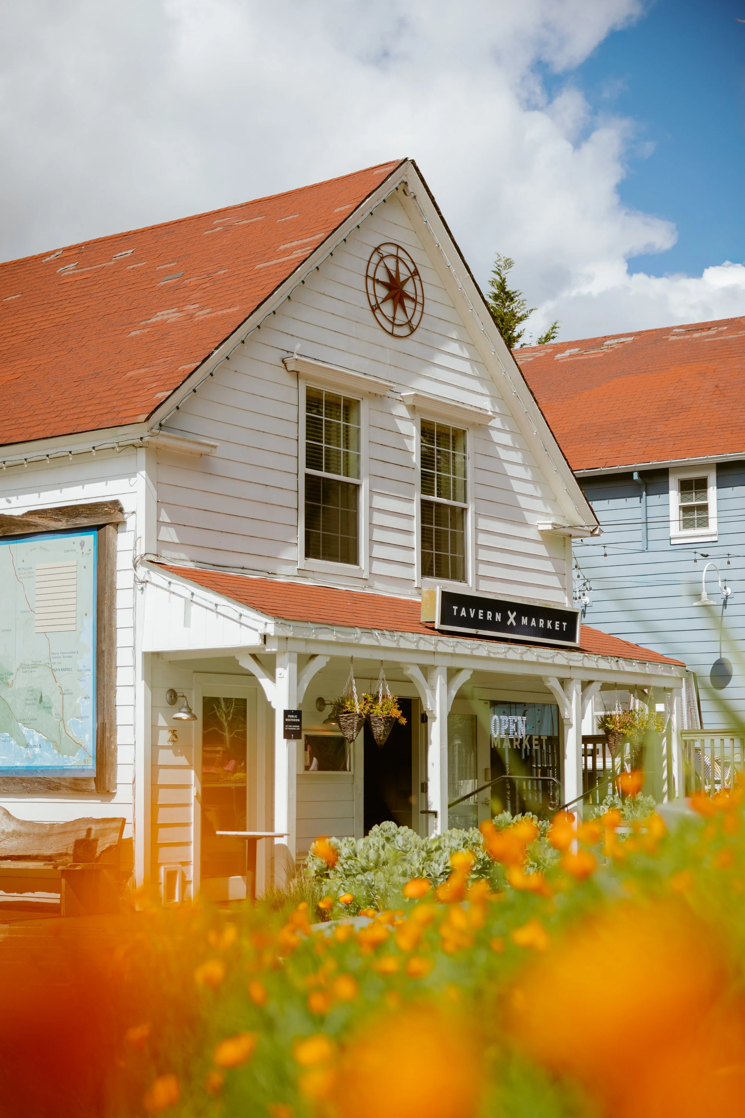 White wooden building with a red-tiled roof and a clock on the gable, storefront reads 'Tavern X Market,' surrounded by flowering plants and string lights, under a partly cloudy sky.