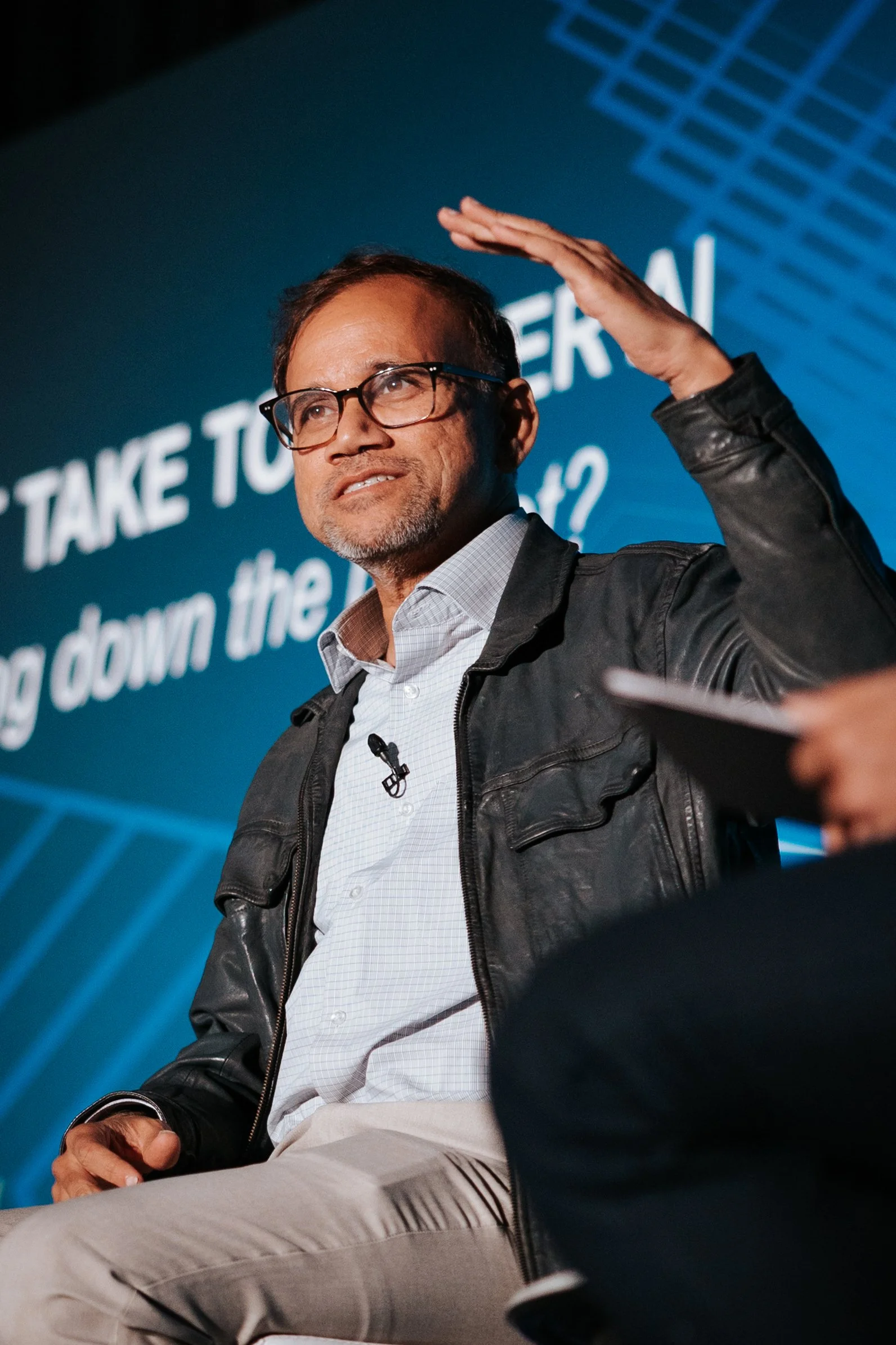 A man wearing glasses, a white checked shirt, and a black leather jacket seated on a panel, raising his right hand near his head while speaking at an event with a screen in the background.