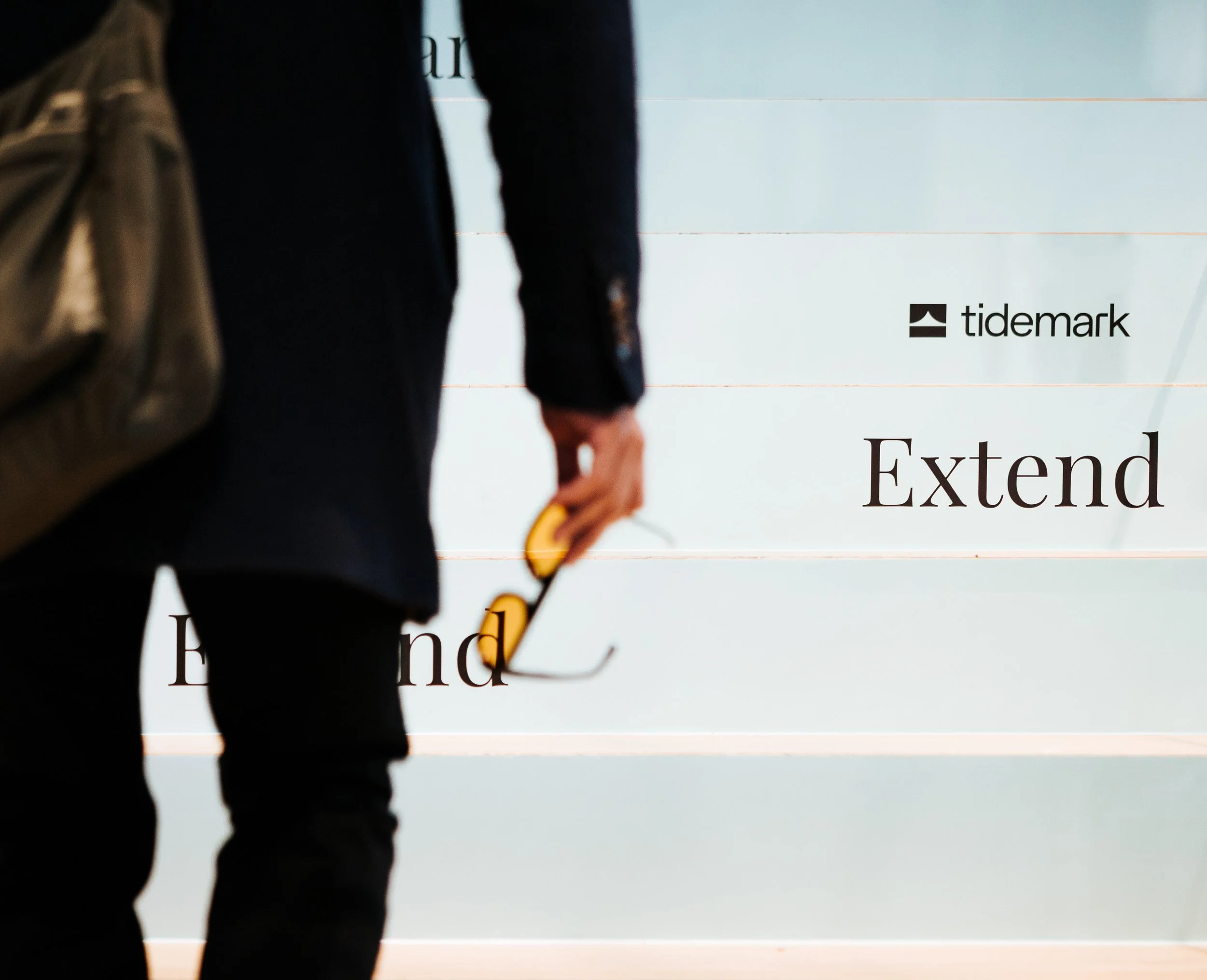 A person in a black suit holding sunglasses and a yellow device, standing in front of a wall with text that reads "Extend" and brand logo "tidemark."