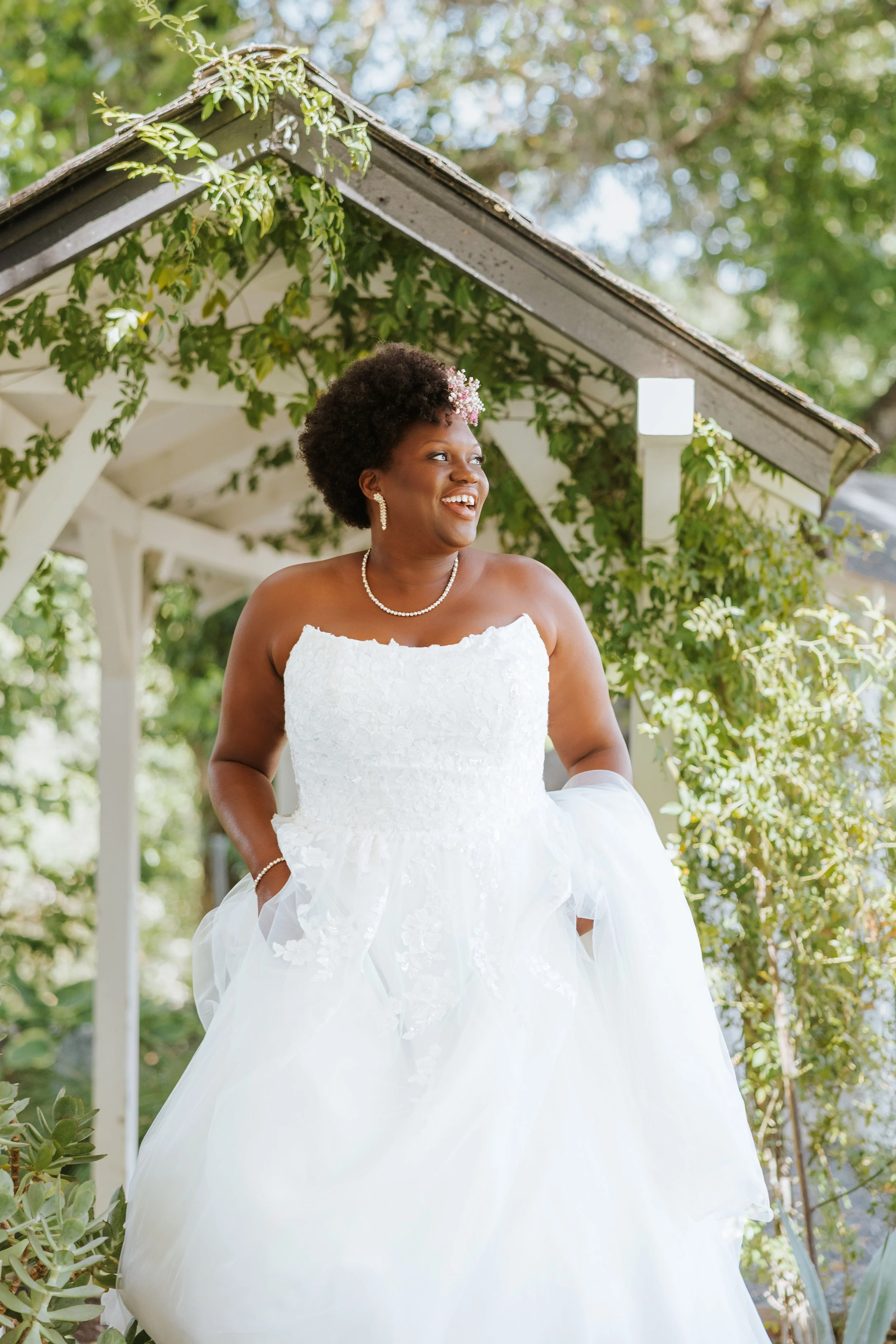 A smiling woman in a white wedding dress with pearl jewelry, standing outdoors under a wooden arbor covered with greenery and flowers.