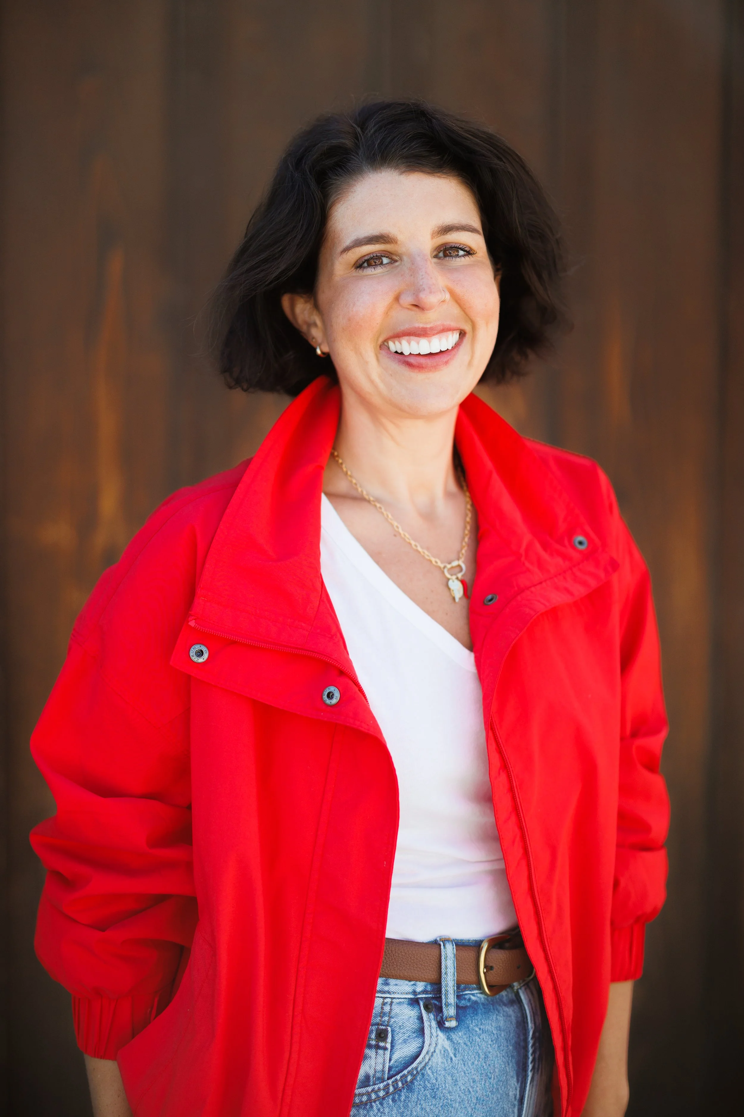 A woman with short dark hair, wearing a red jacket over a white shirt, smiling outdoors with a wooden background.