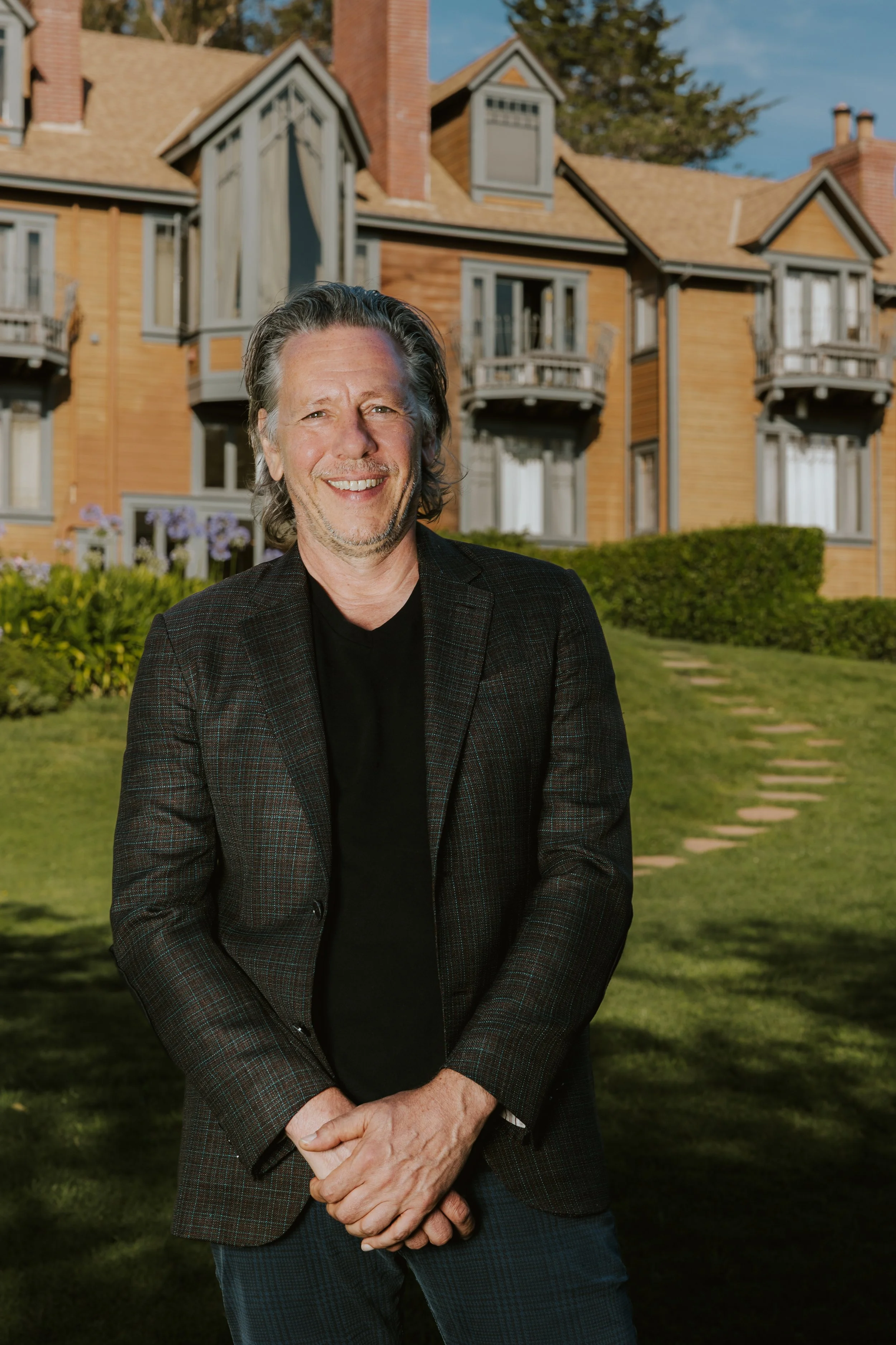 Smiling man in a black blazer with a plaid pattern, standing outdoors in front of a multi-story wooden house with balconies and gabled roofs, lush green lawn, and flowering plants, under a partly cloudy sky.