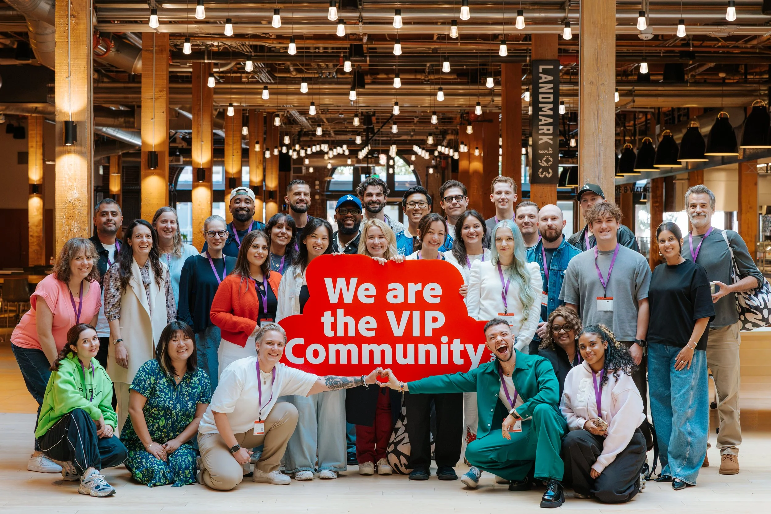 Group of diverse young adults and a few older people gathered indoors, smiling and posing for a photo with a red sign that says 'We are the VIP Community.' The setting appears to be a modern, well-lit space with wooden pillars, overhead lights, and a