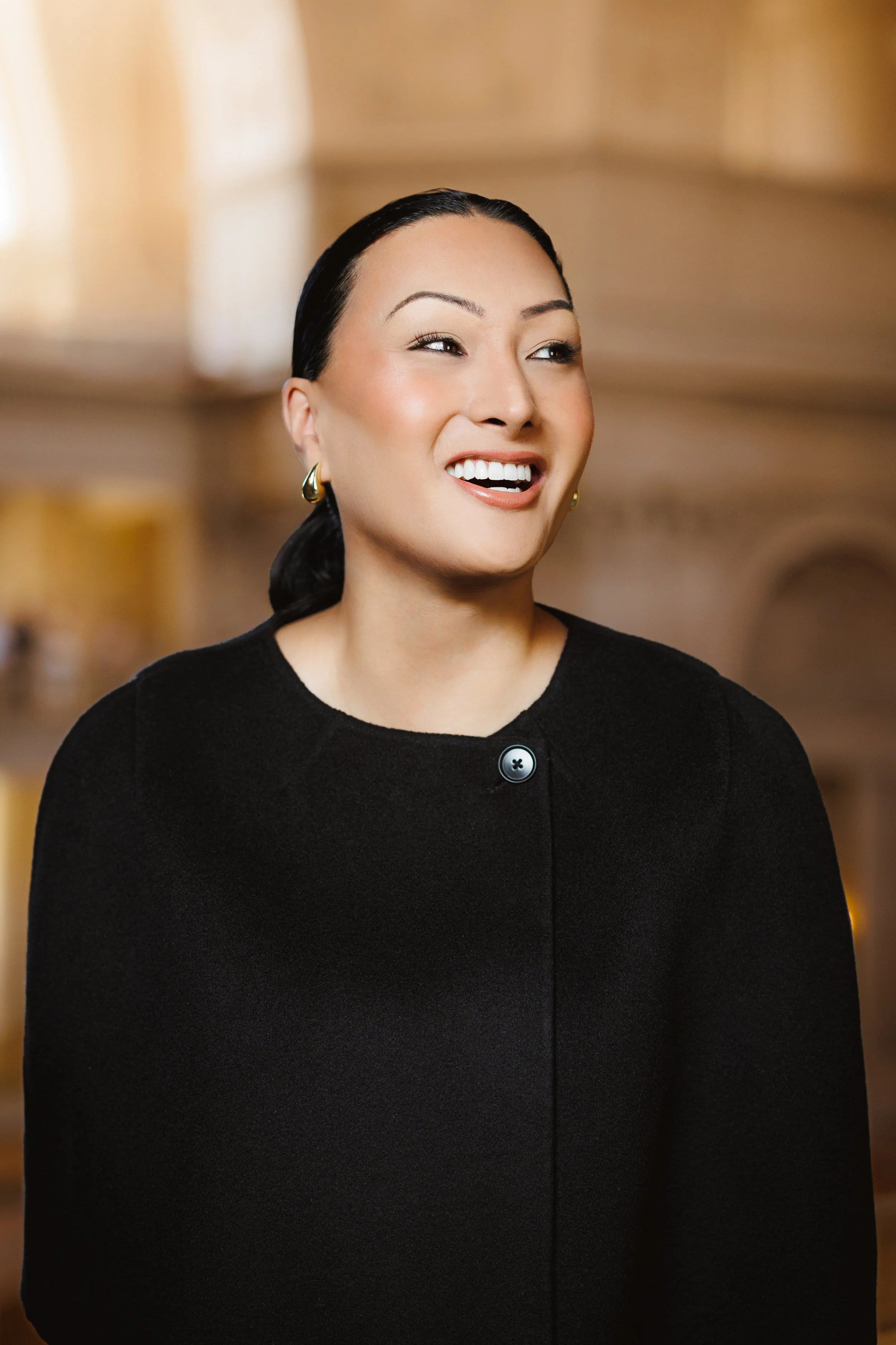 Close-up of a smiling woman with black hair, wearing a black top and gold earrings, in an indoor setting with warm lighting.