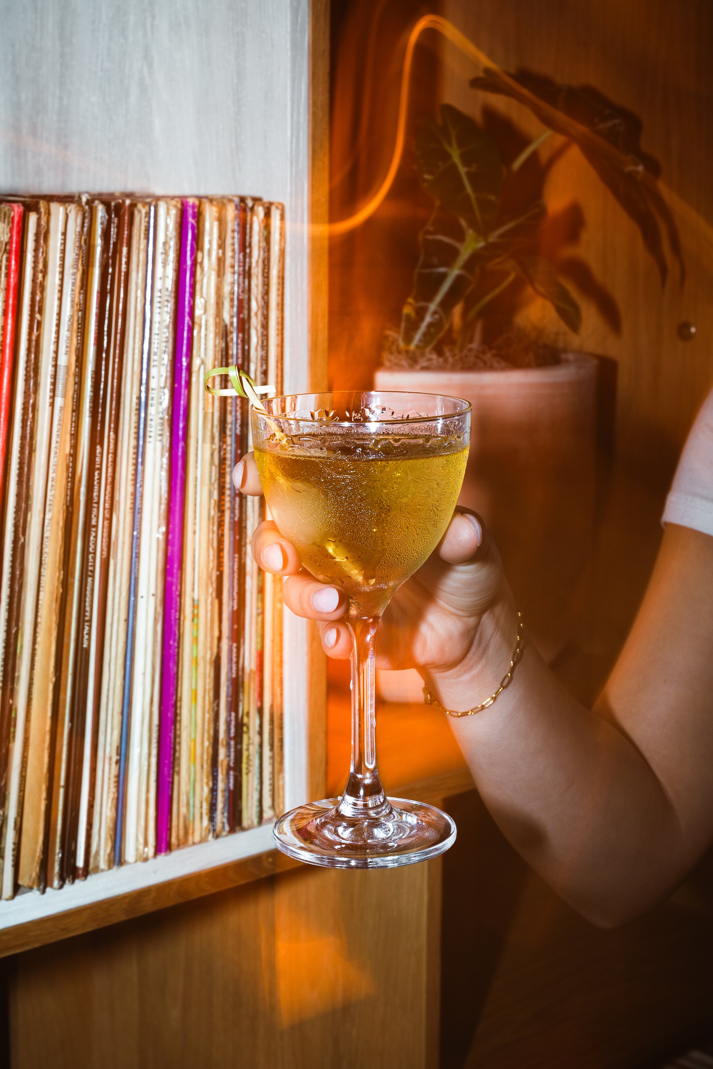 Person holding a cocktail glass with a yellow drink, standing next to a shelf of vinyl records and a potted plant in a warmly lit room.