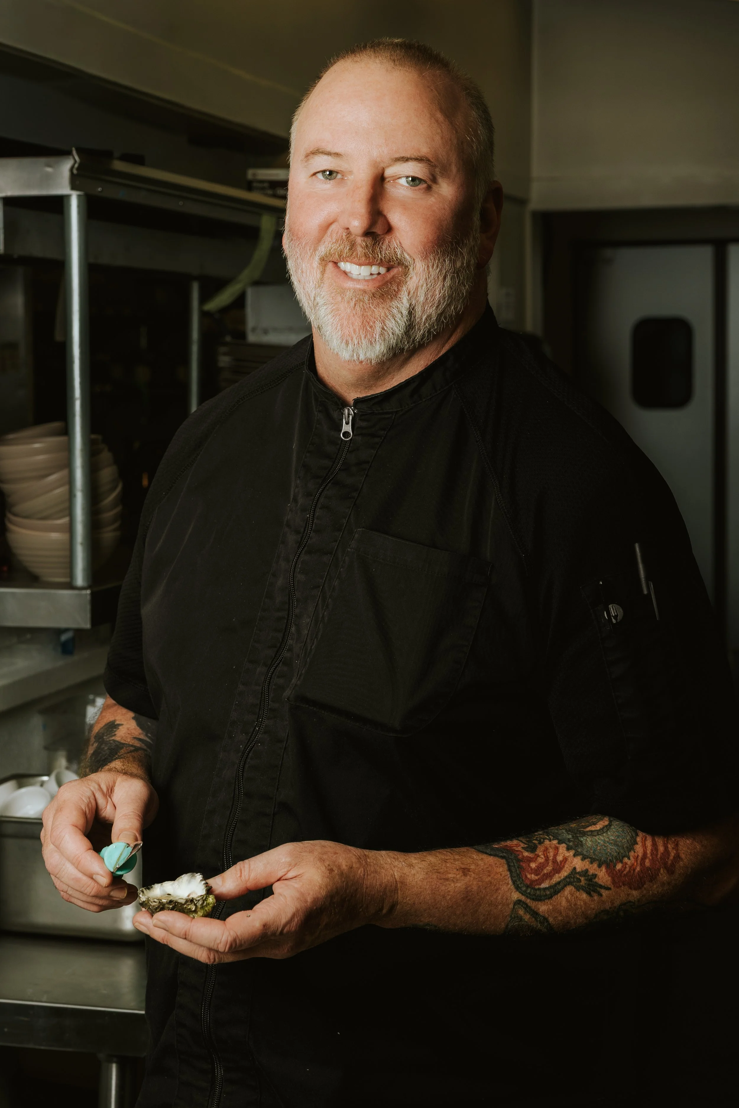 Man with a beard and tattoos in a kitchen, wearing a black chef's jacket, holds a shellfish topped with sauce and a spoon.