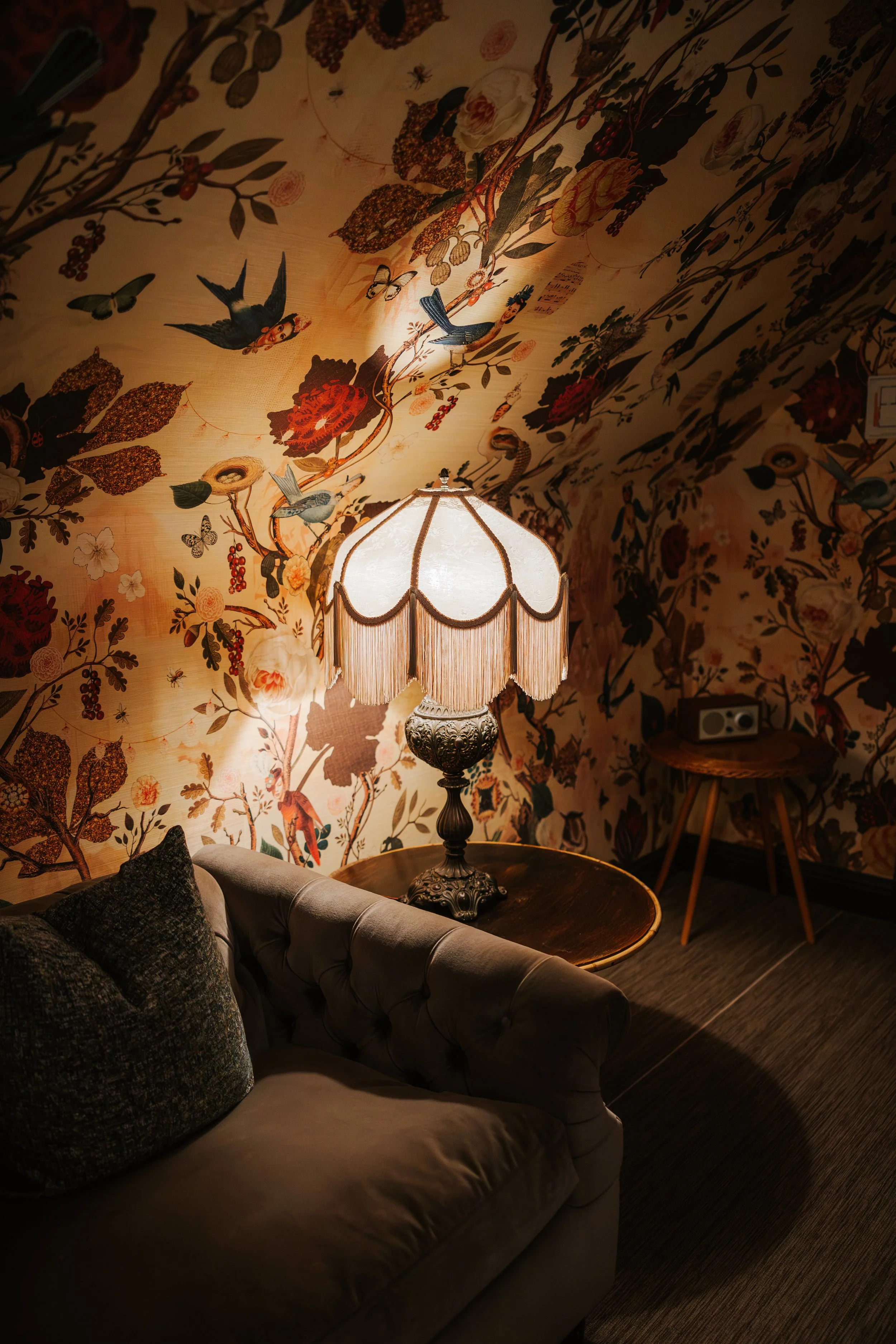 A cozy living room with floral wallpaper, a vintage-style lamp with fringe, a dark sofa with textured cushions, a small wooden side table, and wood flooring.