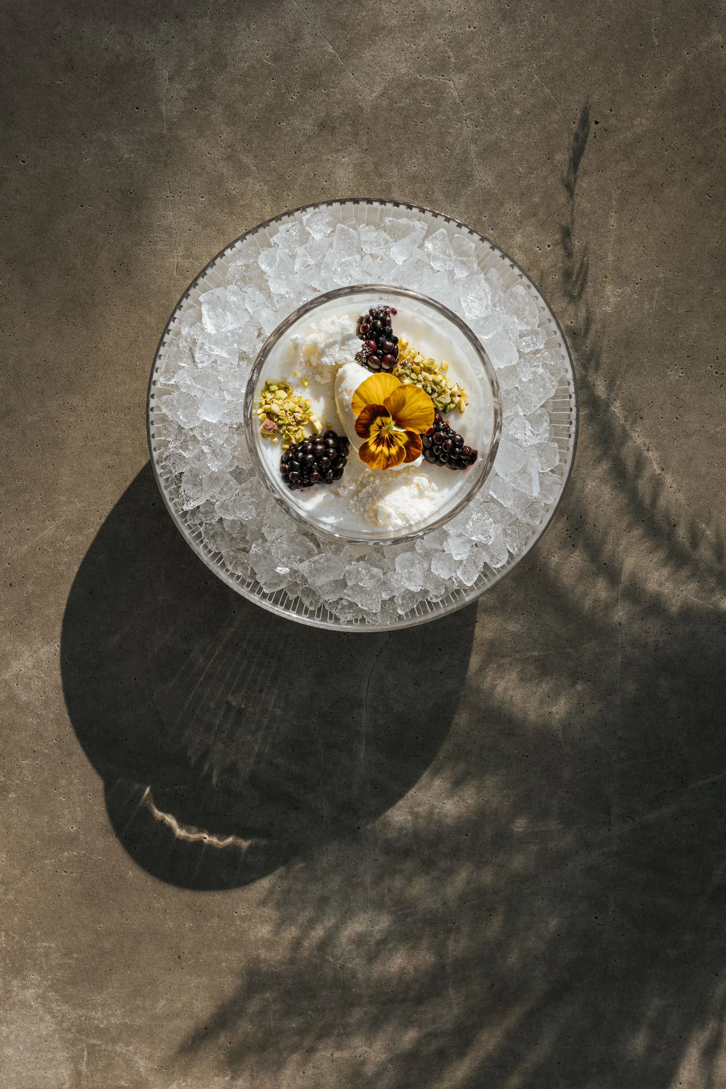 A glass bowl of vanilla ice cream topped with blackberries, chopped pistachios, whipped cream, a yellow edible flower, and a brown flower petal, placed on a larger glass plate filled with ice cubes on a concrete surface.