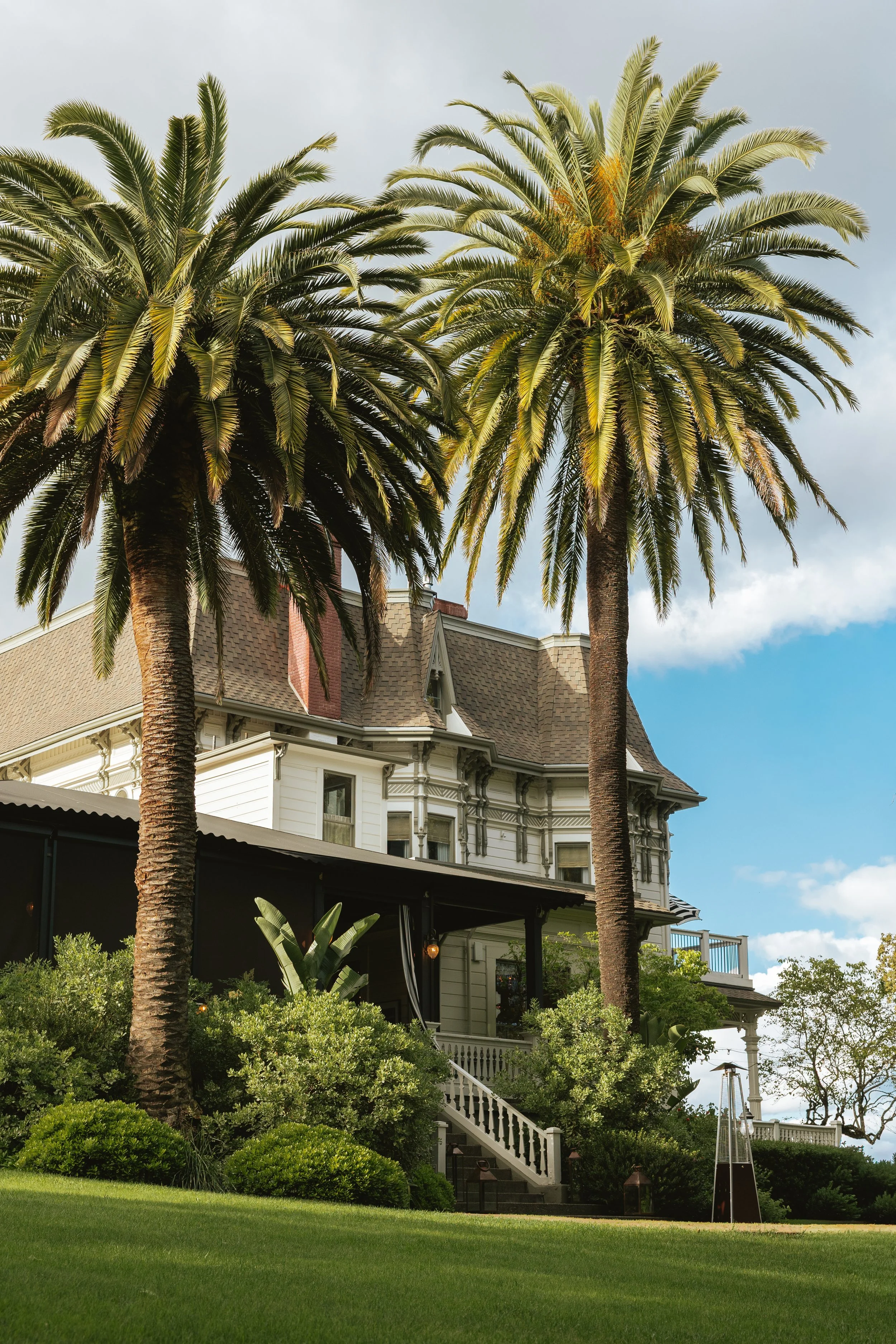 A Victorian-style house with pastel yellow siding, ornate trim, and a steep roof, surrounded by lush greenery including palm trees, shrubs, and a well-maintained lawn, with a cloudy sky in the background.