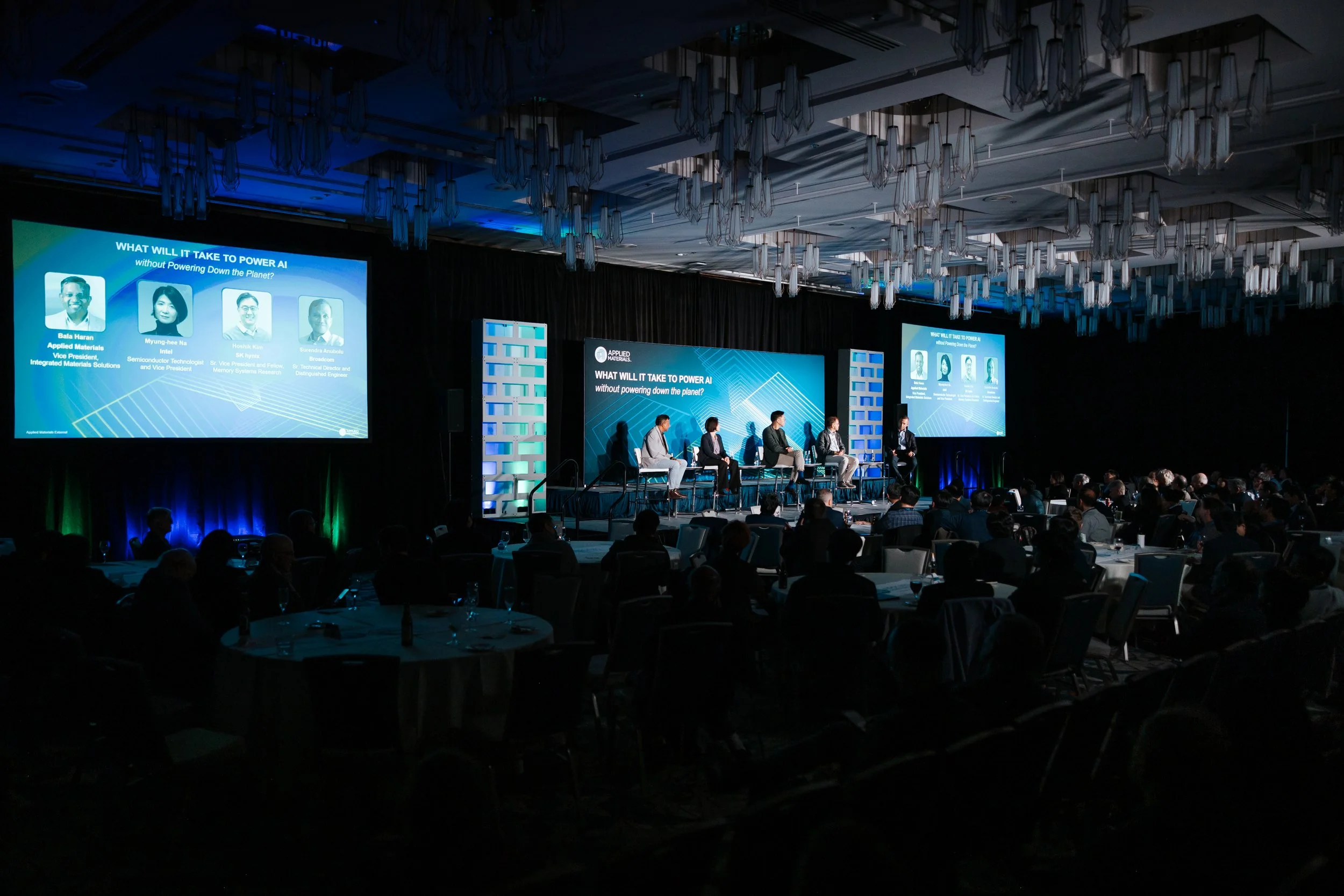 A conference or panel discussion taking place in a darkened room with a large audience, five panelists seated on stage, and two large screens displaying the topic 'What will it take to power AI without powering down the planet?'.