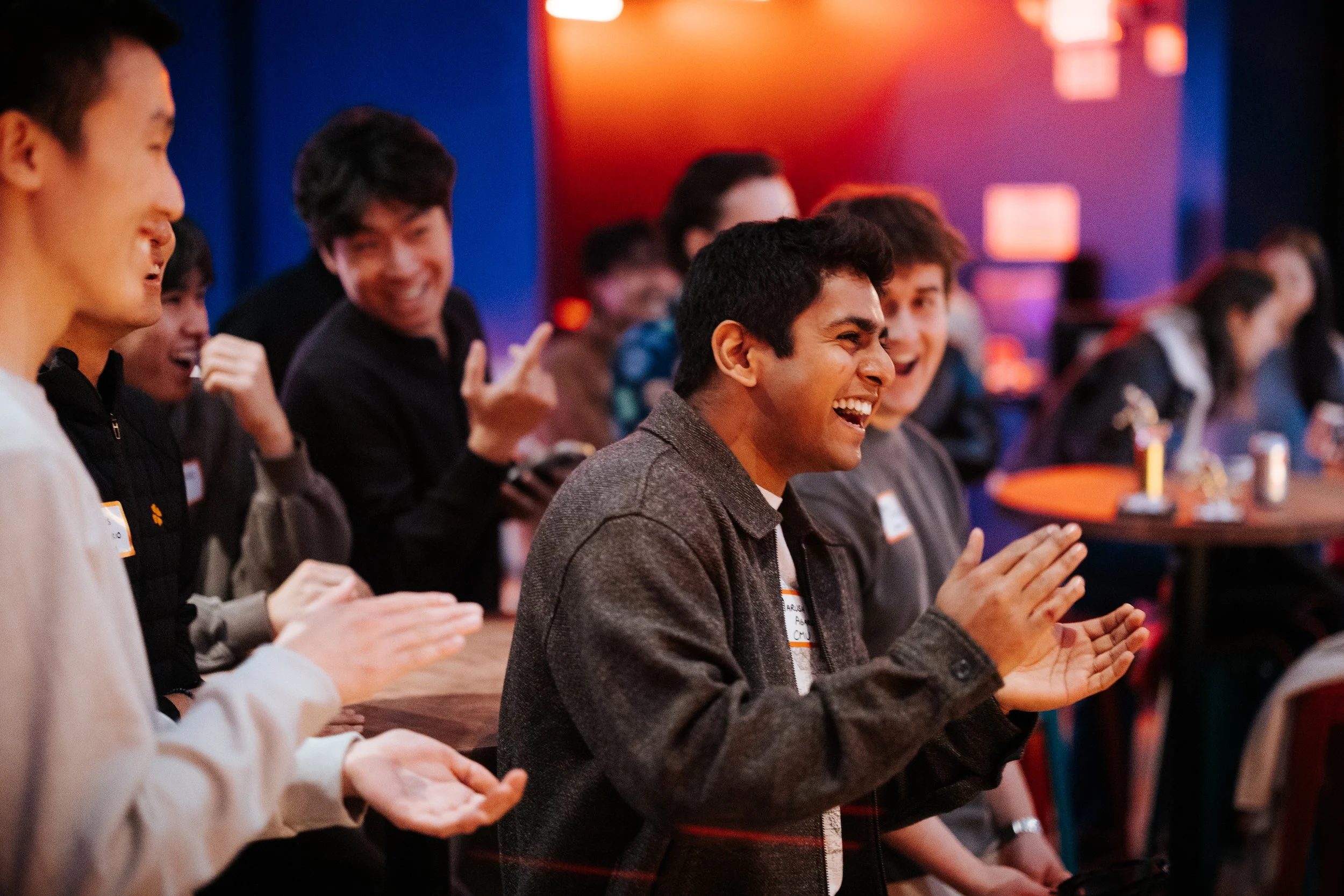 Group of young people enjoying a social event, they are smiling, clapping, and laughing in a lively indoor setting with colorful lighting.