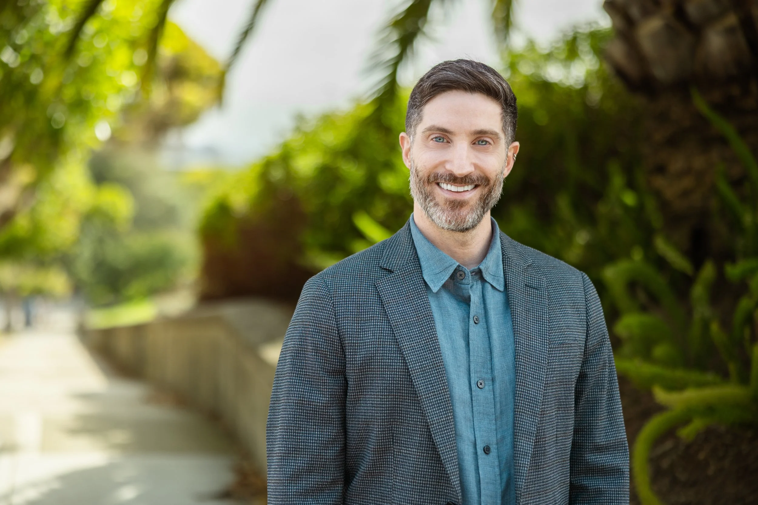 Portrait of a smiling man with dark hair and a beard, wearing a blazer and blue shirt, standing outdoors with green trees in the background.