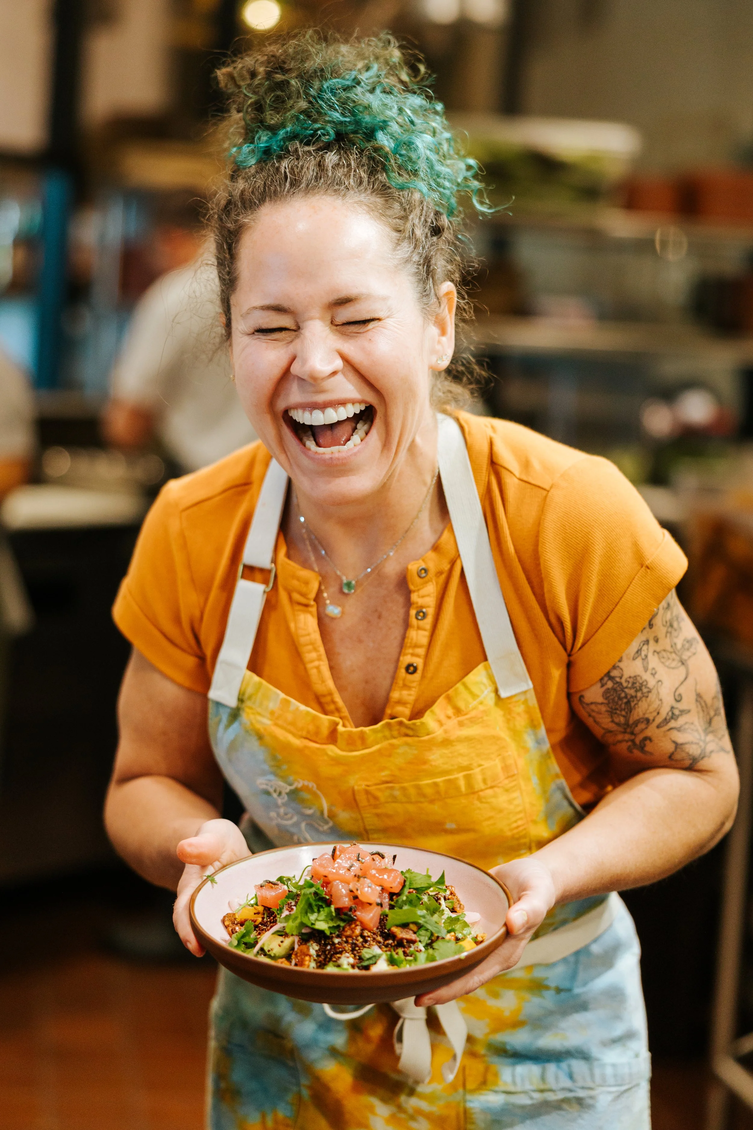 A woman with curly hair, wearing an orange shirt and apron, holding a bowl of salad in a kitchen, smiling and laughing.
