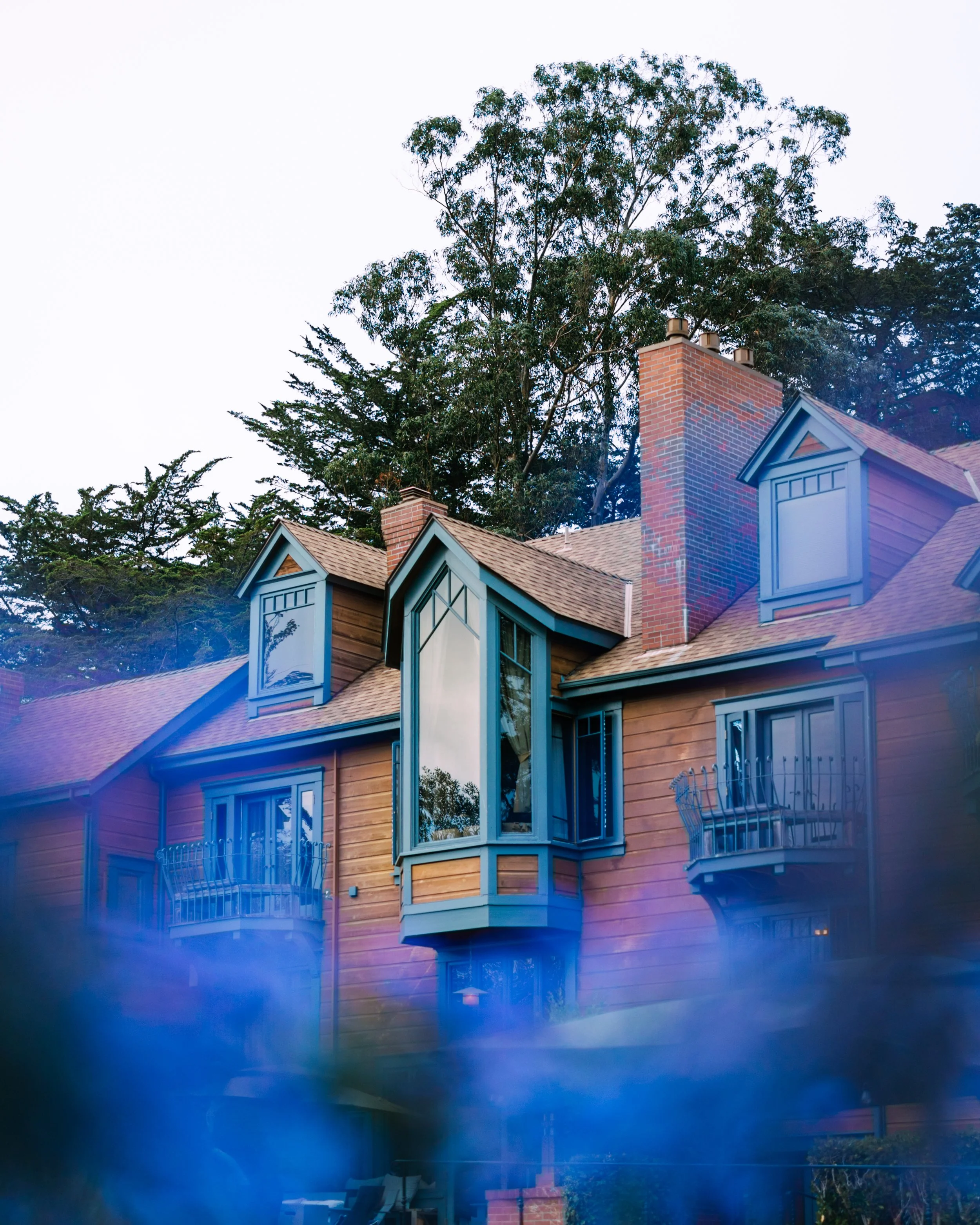 Olema house with a brick chimney, blue trim, and a balcony, surrounded by tall trees in the background, under a clear sky.