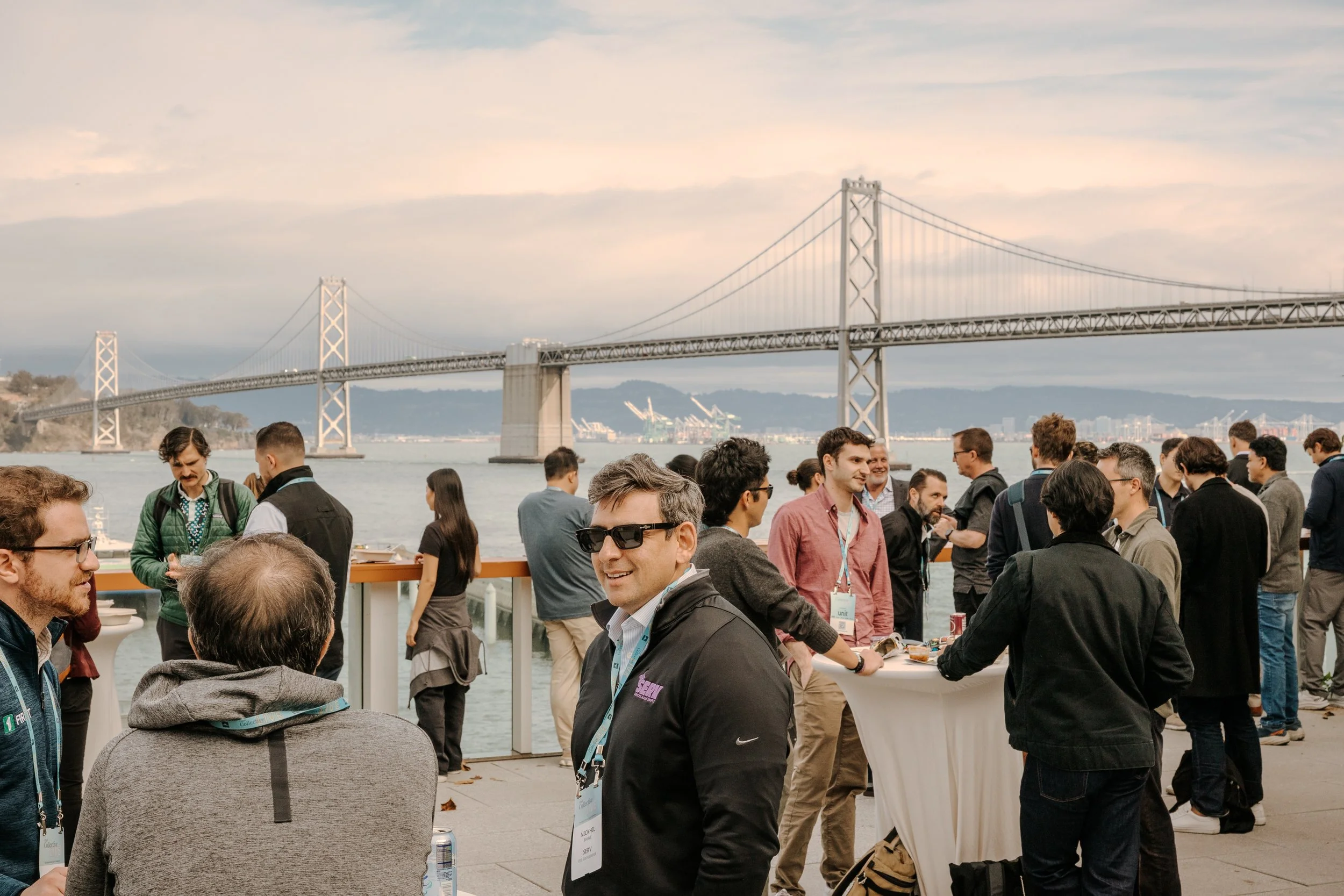 Group of people attending an outdoor event on a deck with a view of a large bridge and water in the background during daytime.