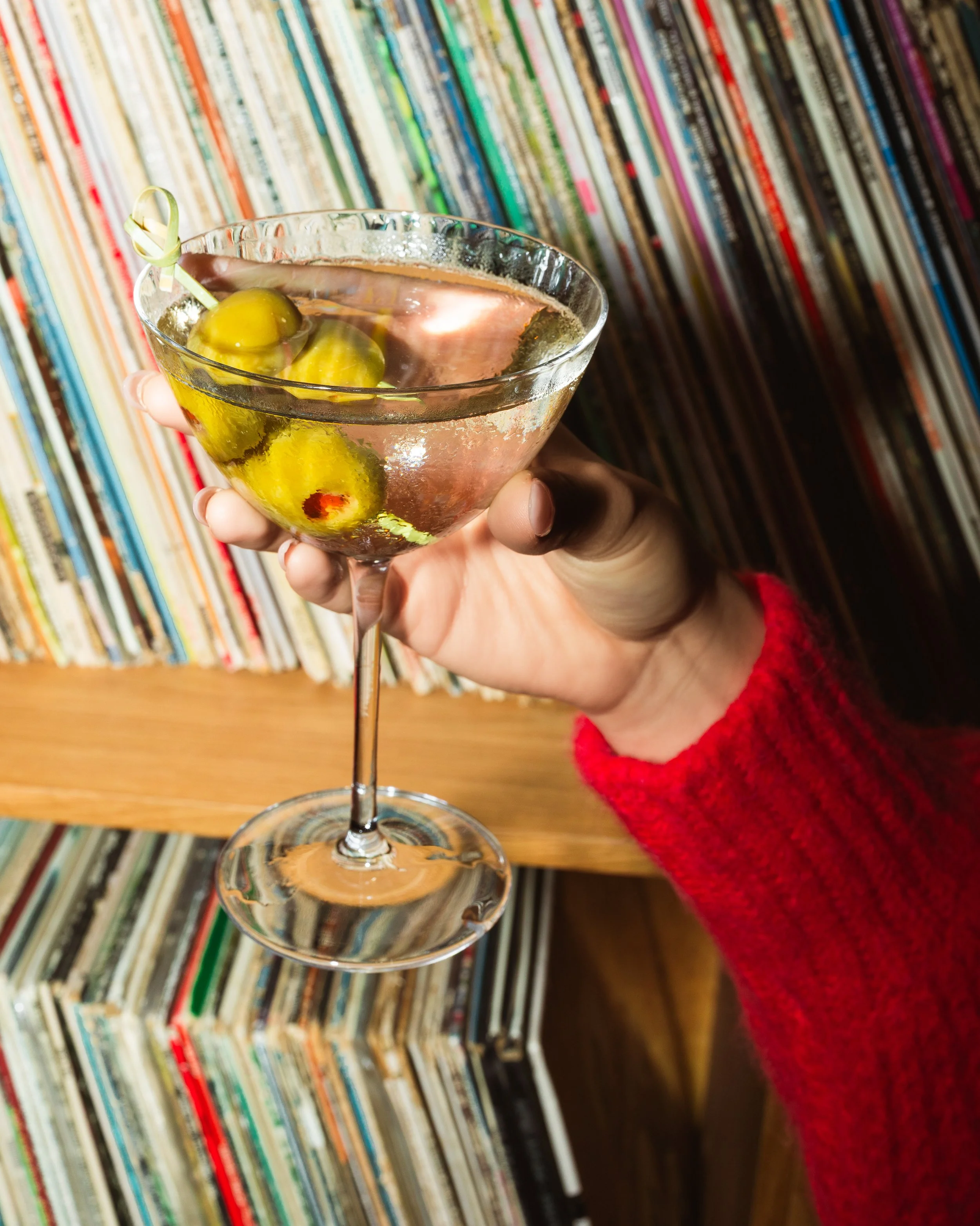 A hand holding a cocktail glass filled with a pink liquid and green olives, with a skewer, against a background of vinyl records.