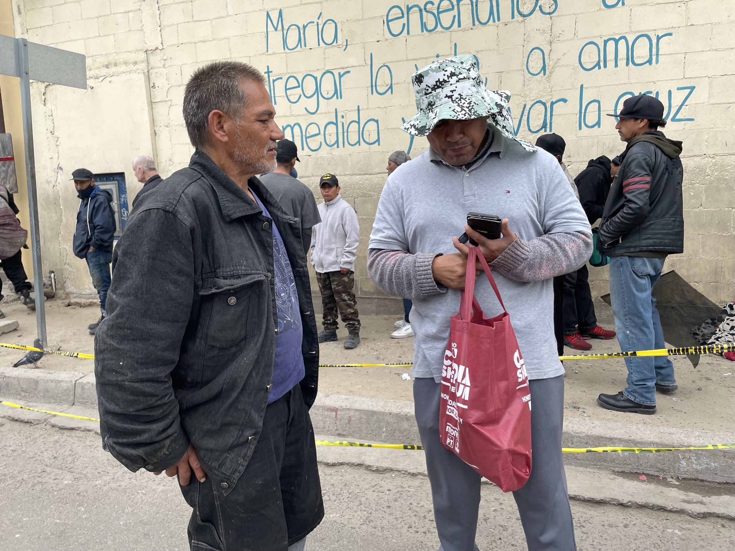 Group of people gathered on a street near a wall with Spanish text, caution tape, and a man holding a smartphone and a red bag.