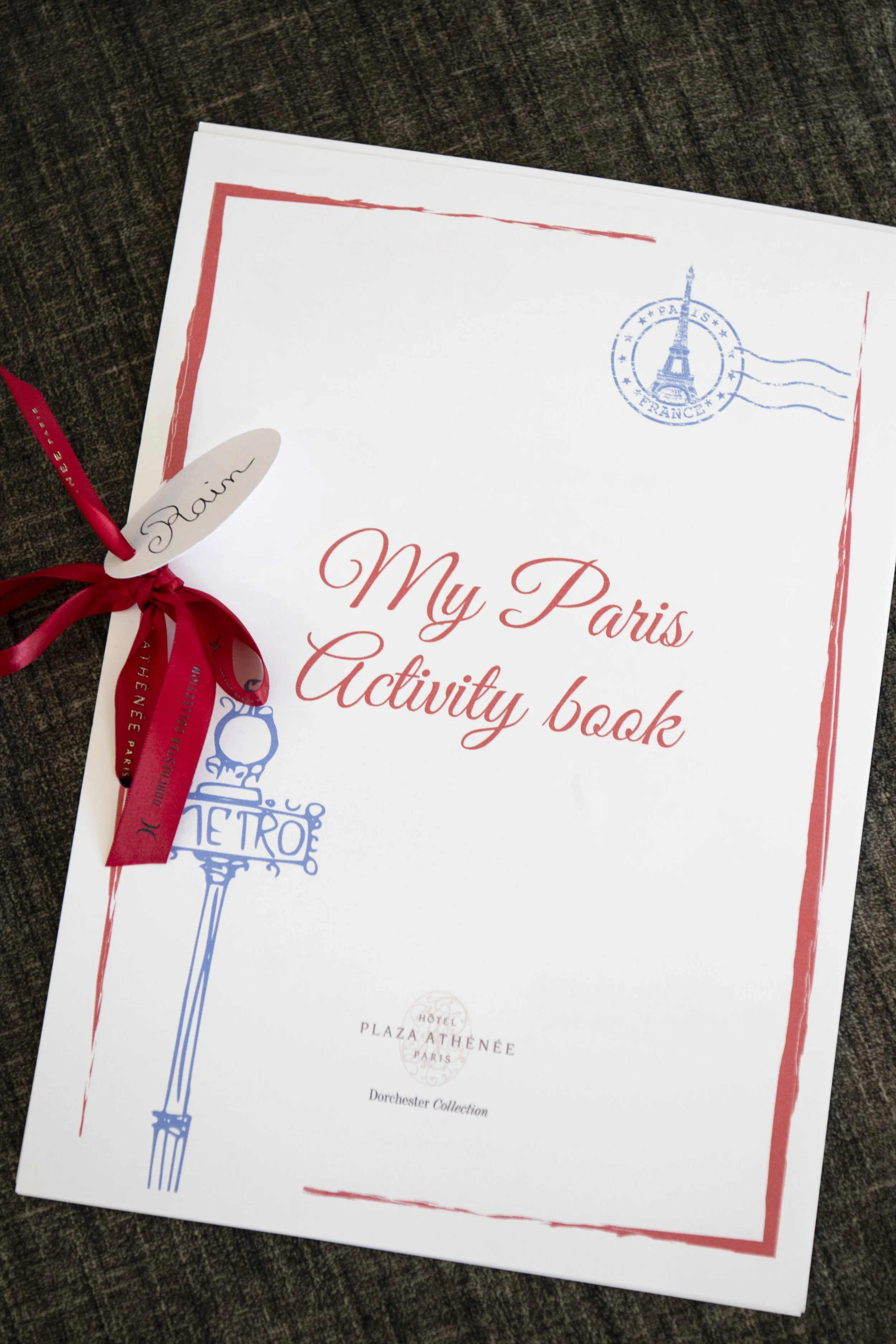 A white booklet titled "My Paris Activity Book" with a blue Eiffel Tower illustration, a red ribbon, and a postal stamp featuring the Eiffel Tower on a dark fabric surface.