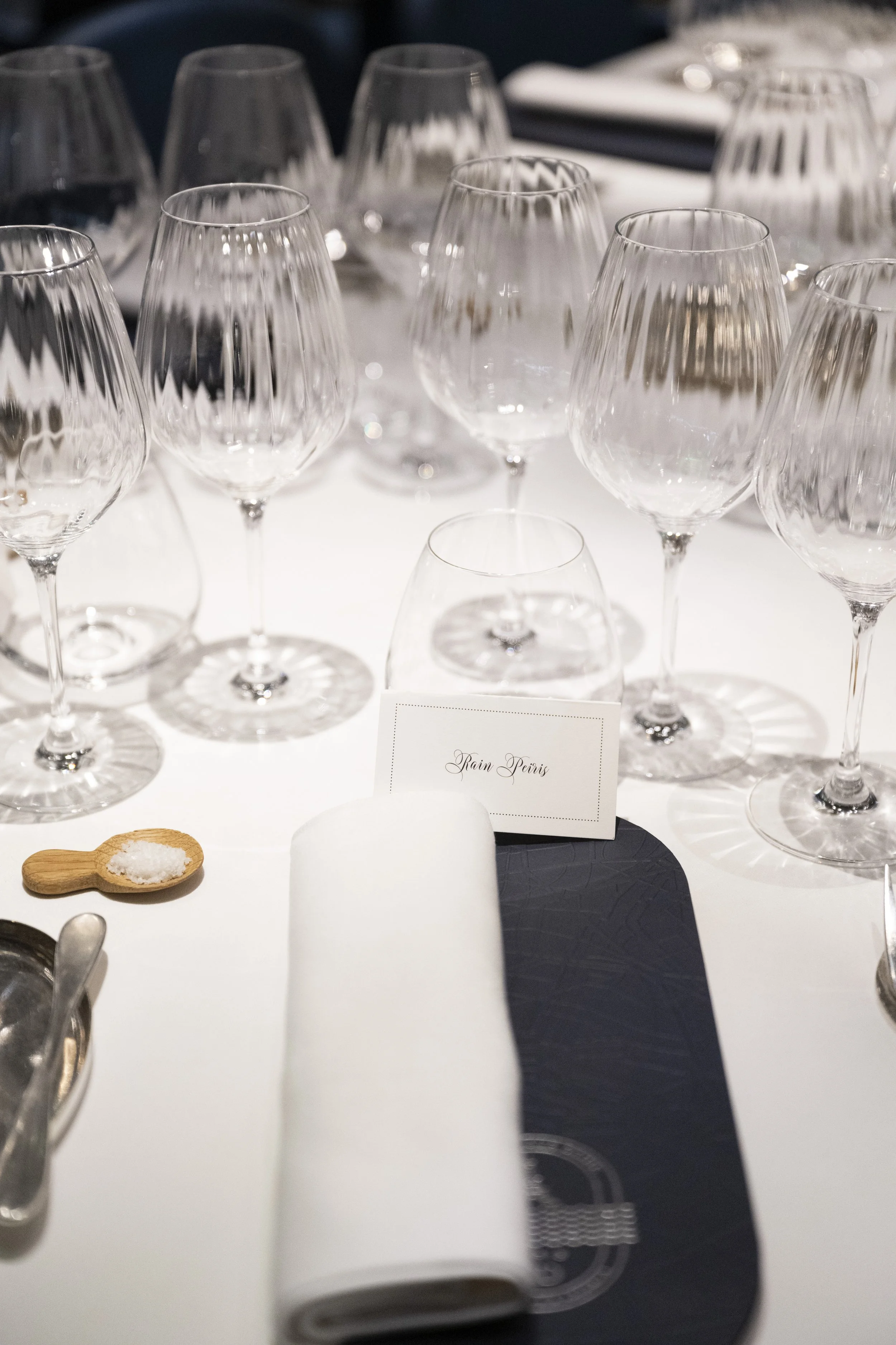 Multiple empty wine glasses on a white table, with a small white card in the center table reading 'Train Paris,' a small wooden spoon with salt, and some black tableware. The background is blurred.