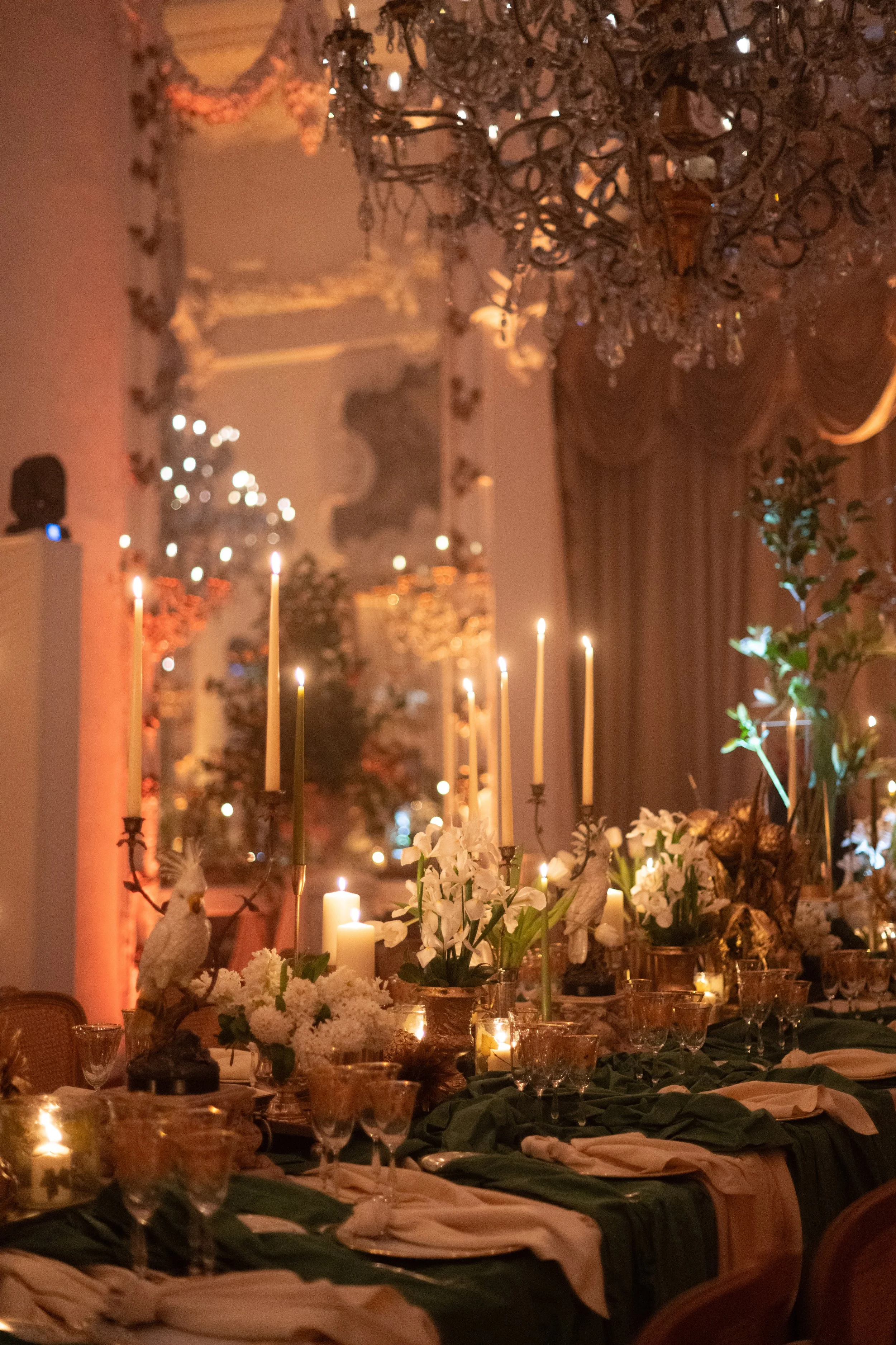 An elegant dining room decorated for a special occasion with lit candles, floral arrangements, and ornate chandeliers. The room features draped curtains and warm lighting.