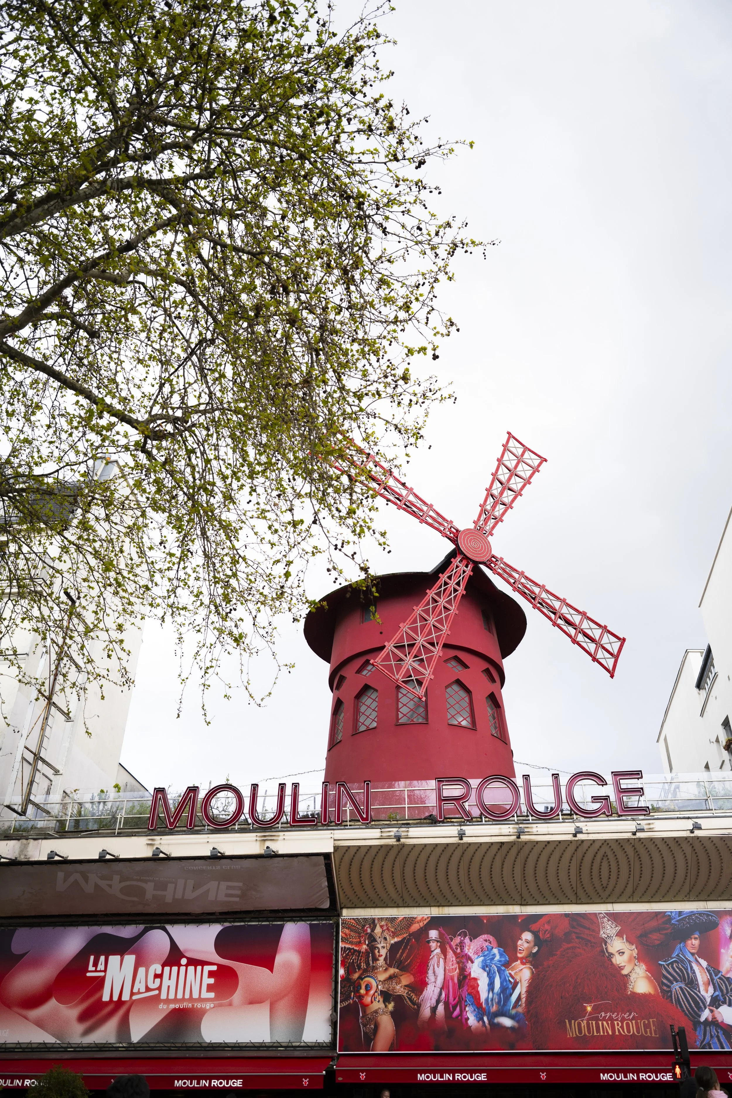 The image shows the Moulin Rouge windmill tower with a red structure and the sign 'Moulin Rouge' below it. Surrounding the windmill are trees and posters, including one for a show called 'La Machine du Moulin Rouge' featuring colorful characters and 