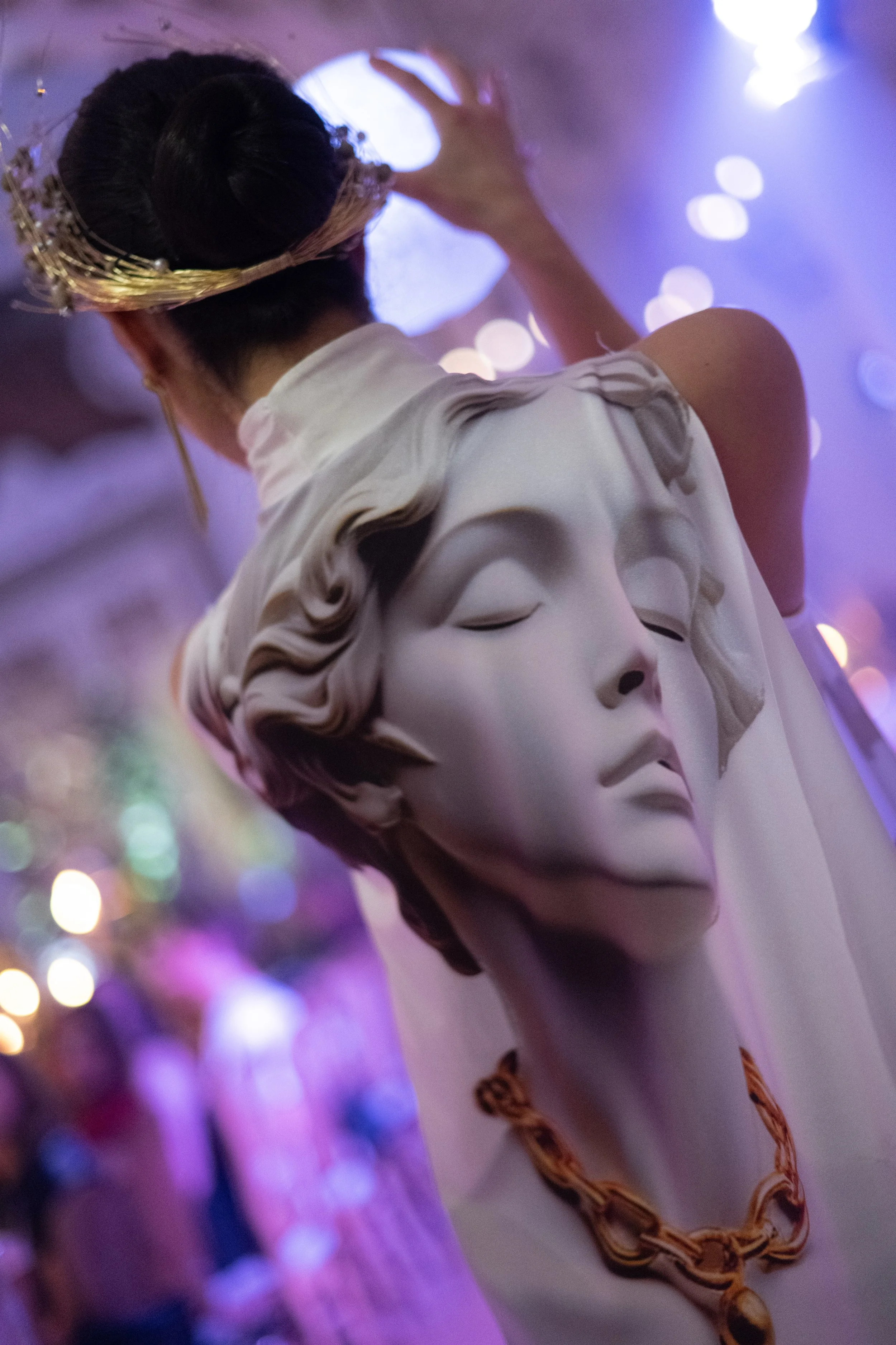 An ornate statue of Venus, the Roman goddess, adorned with a gold crown and jewelry, with a blurred background of purple and white lights.