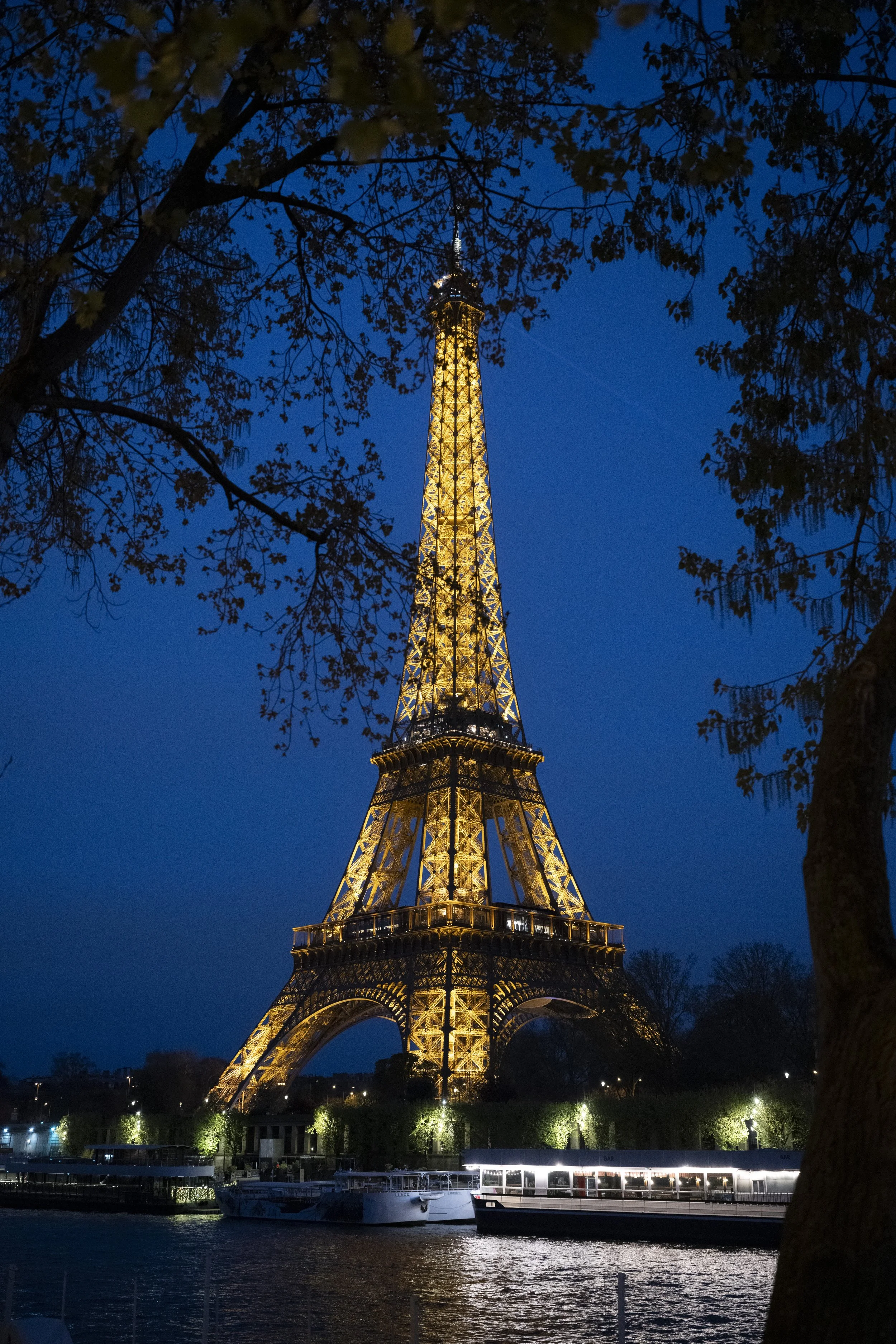The Eiffel Tower illuminated at night, seen through tree branches, near a river with boats and a lit walkway.